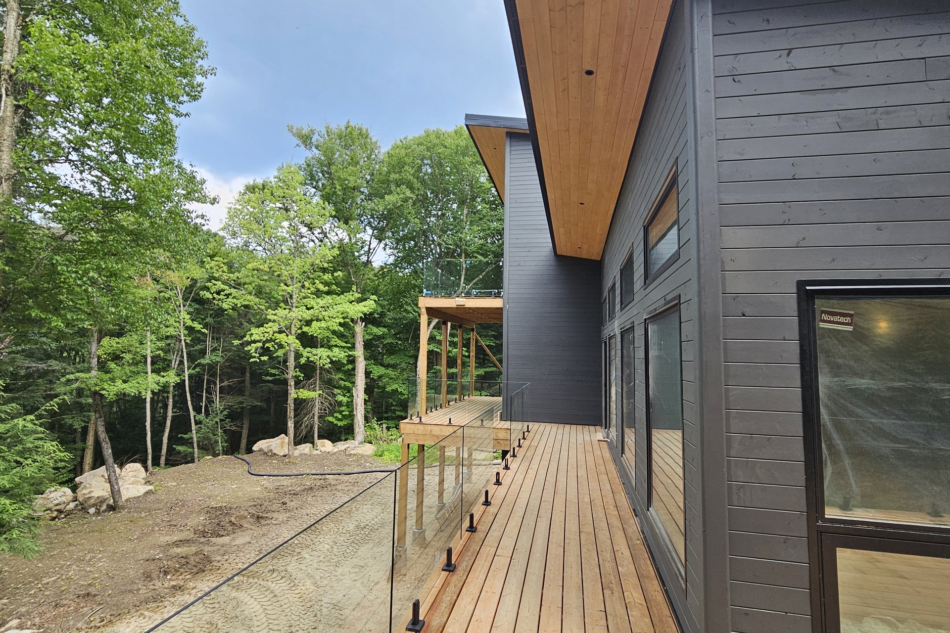 A modern house exterior features dark siding, large windows, and a wooden deck with a glass railing, surrounded by green trees.