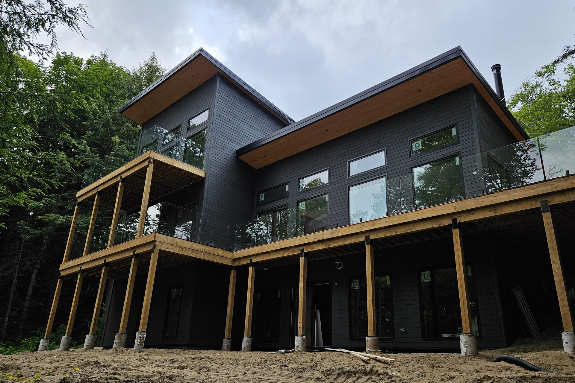 Exterior — Deck & Patio A modern black house with large glass windows and wooden decks is set amidst a lush green forest under a cloudy sky.
