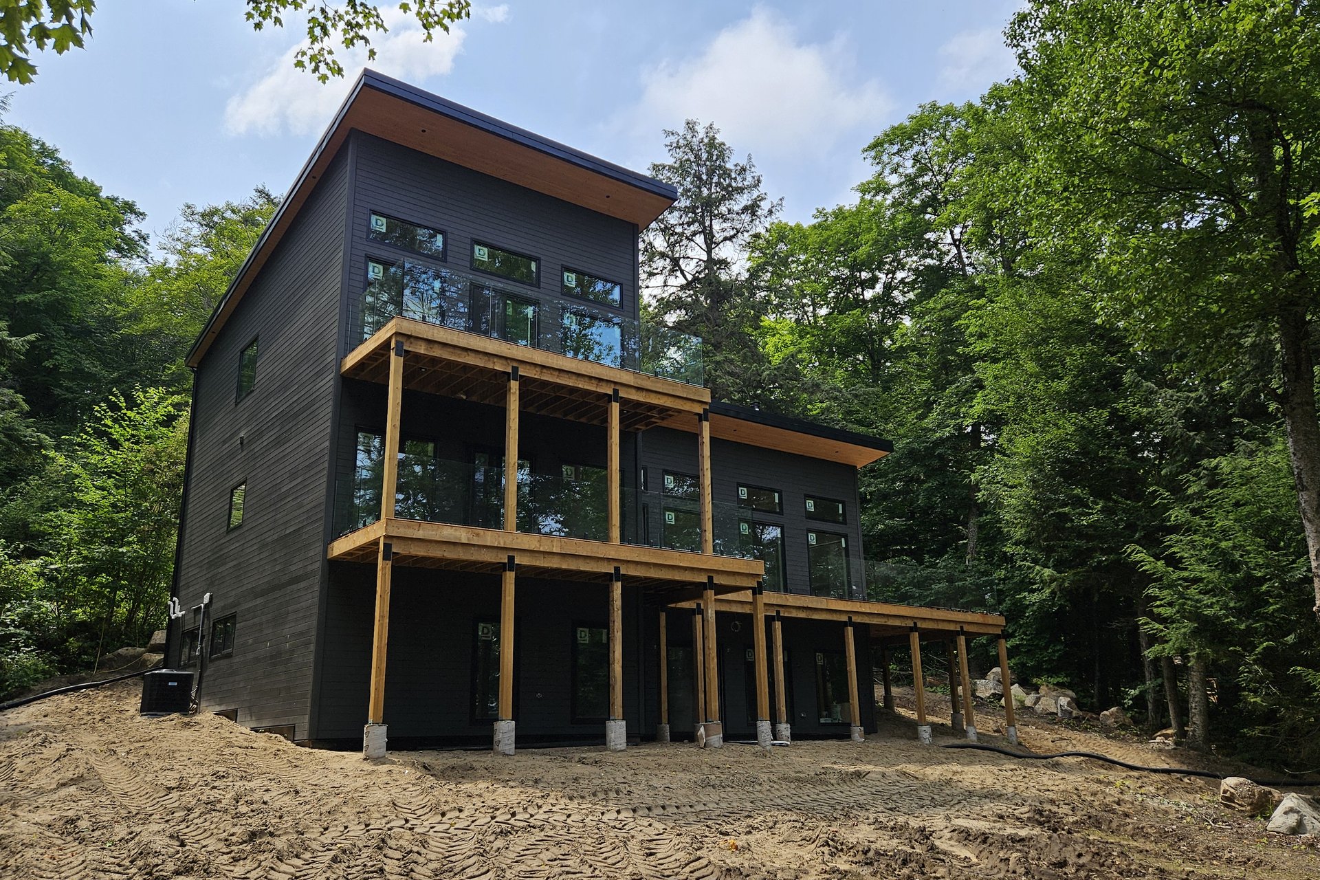 Exterior — Deck & Patio A modern black house with extensive windows and wooden decks is set against a backdrop of dense green trees.