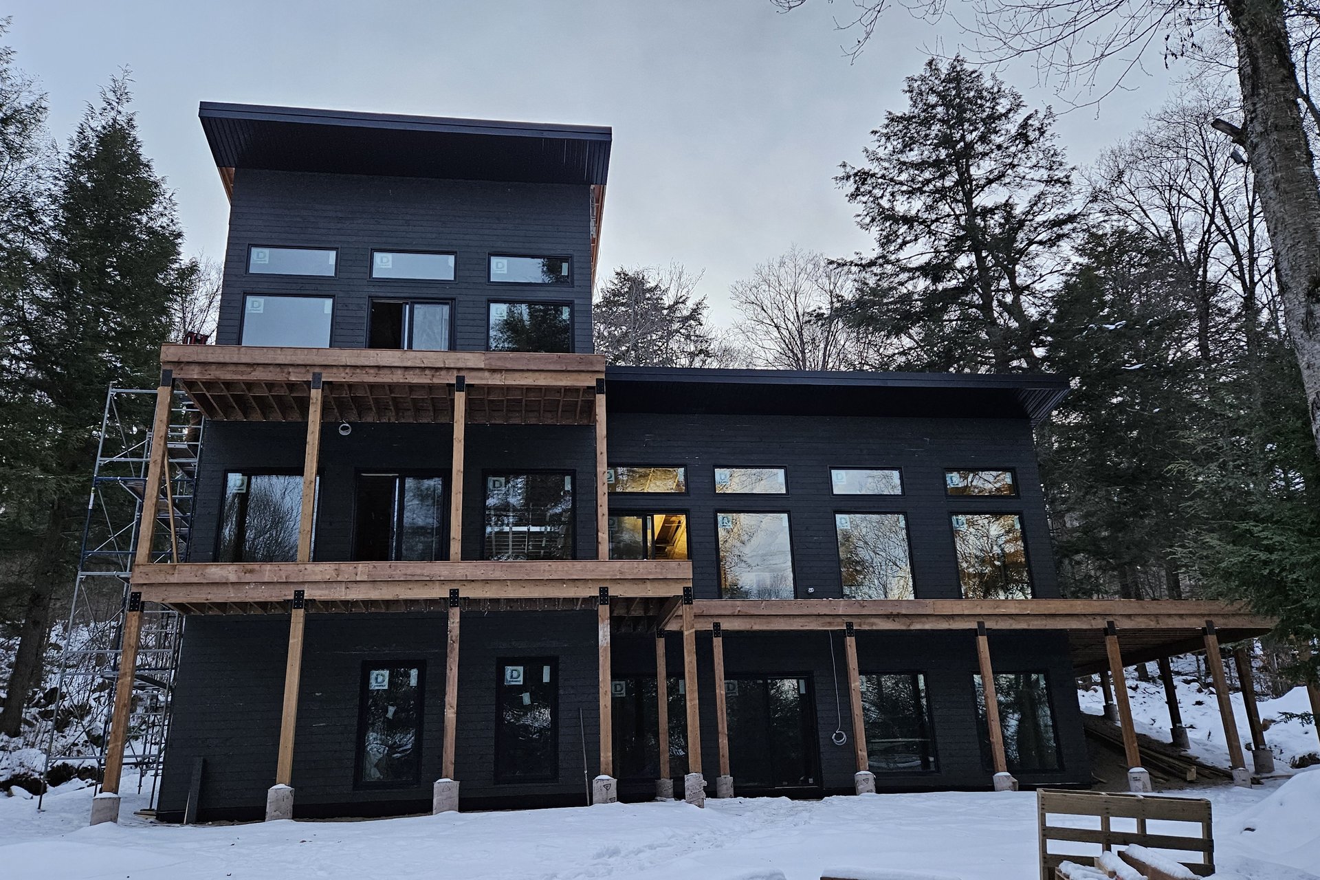 Exterior — Deck & Patio A striking modern dark-sided home with multiple levels of wooden decks is nestled among snowy trees.