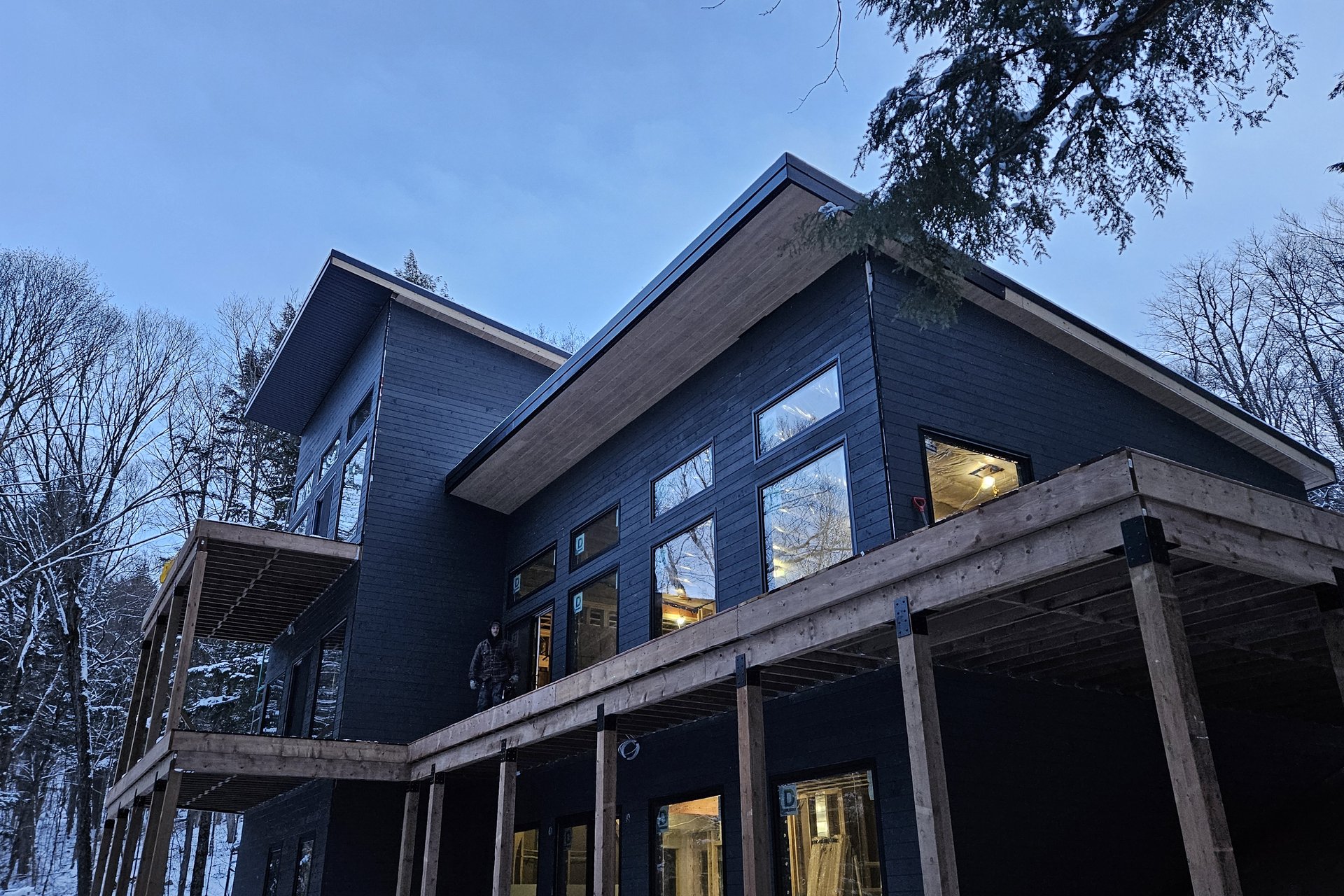 Exterior — Windows & Natural Light A modern dark-colored house with large windows and wooden accents is seen from an exterior angle during dusk in a snowy, wooded area.