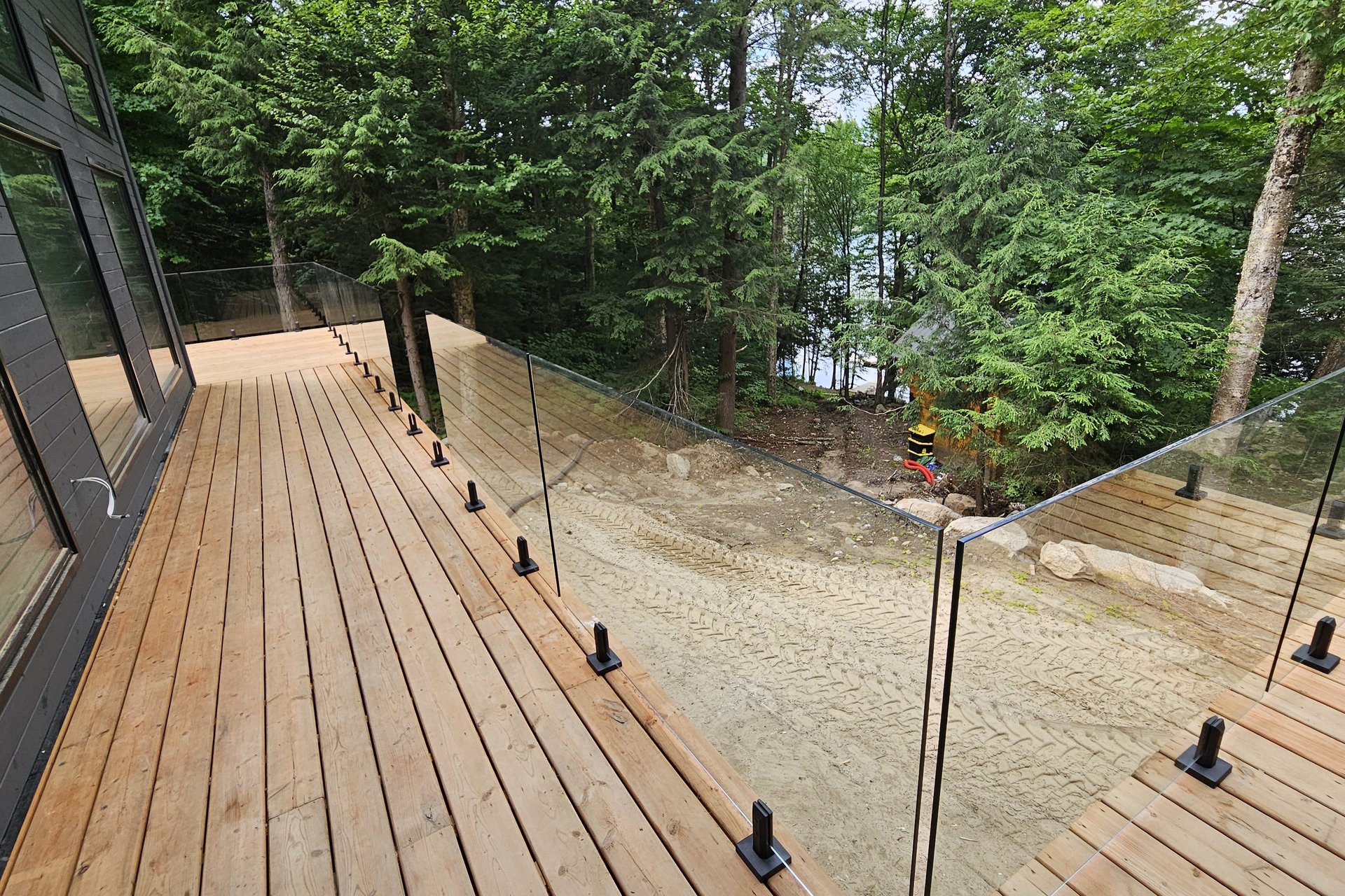 Exterior — Lakefront Deck A newly installed wooden deck with glass railings overlooking a forested area and a body of water.