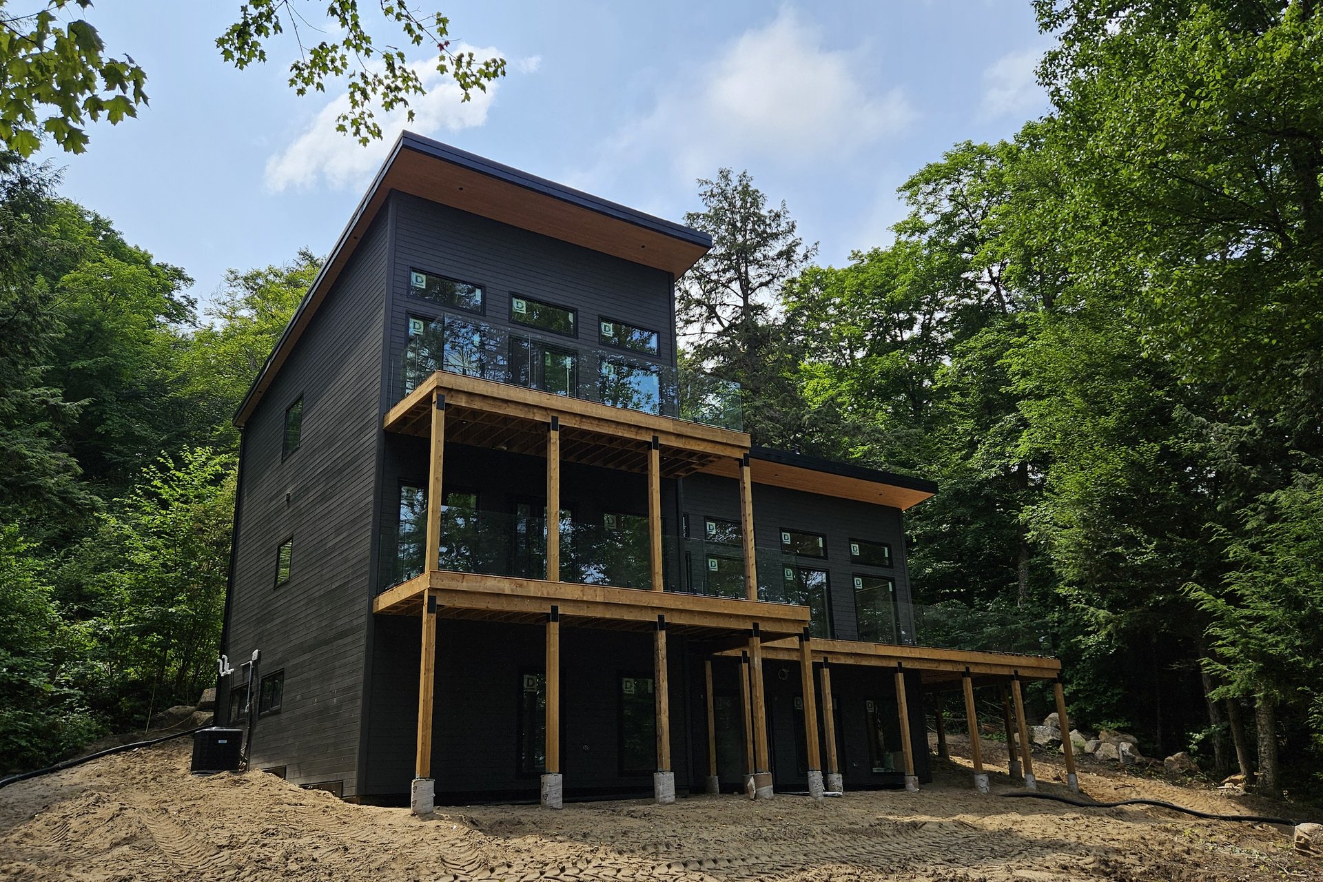 Exterior — Deck & Patio A modern black house with multiple decks and glass railings is nestled in a lush green forest on a sunny day.