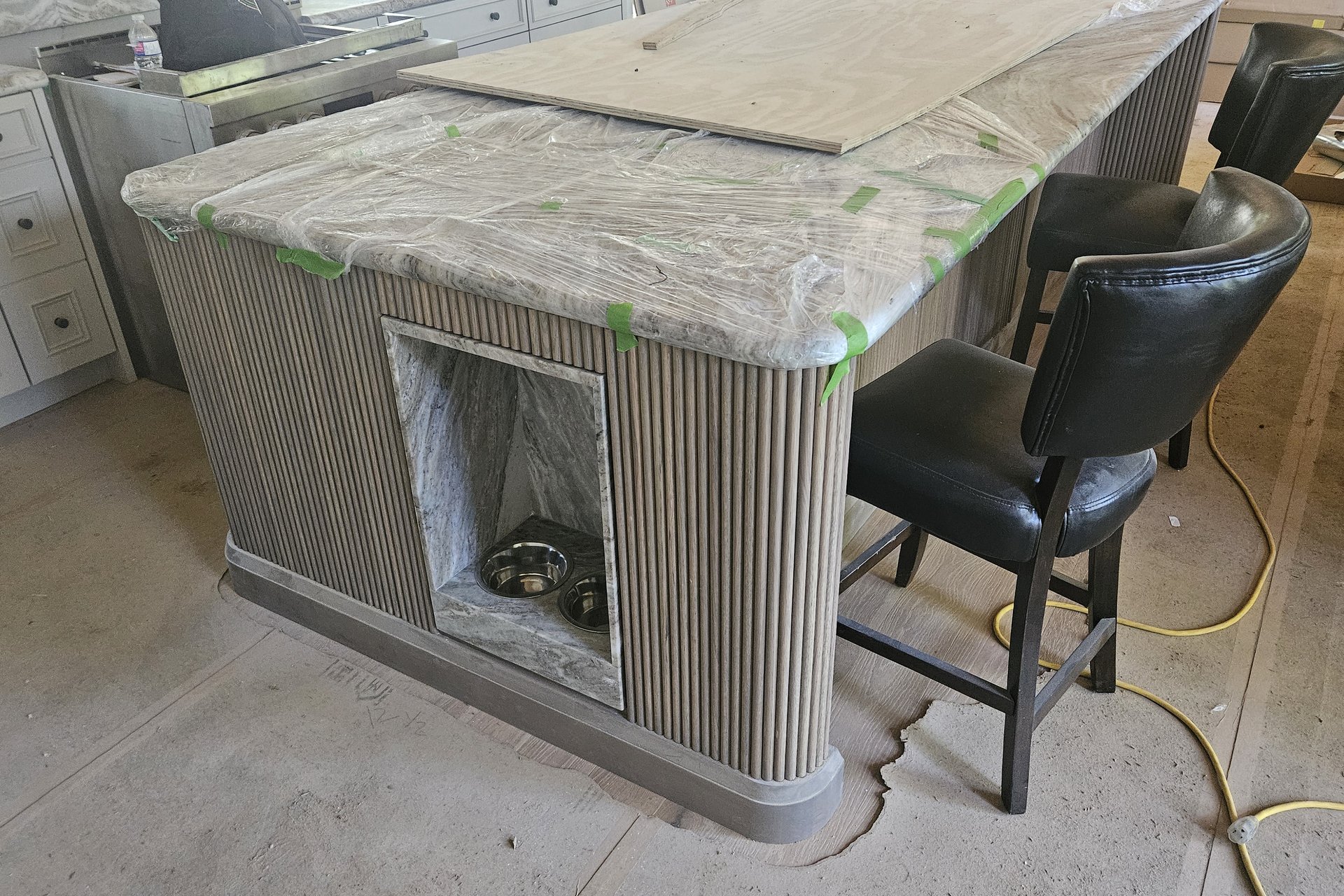 Kitchen — Custom Island A kitchen island with a fluted wooden base and a dog bed cutout is partially covered in plastic, with two bar stools beside it, indicating ongoing interior finishing.