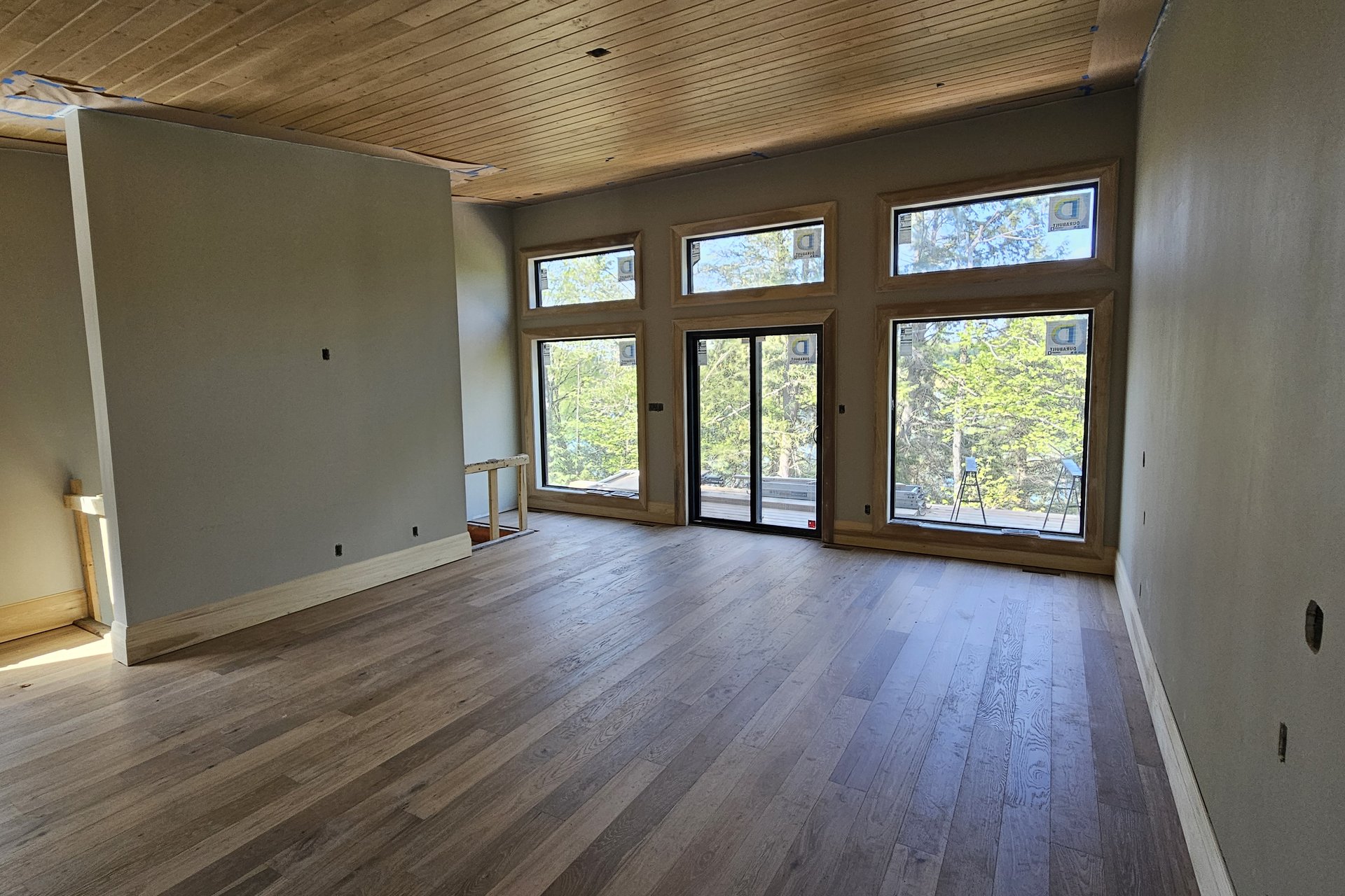 Living Room — Vaulted Wood Ceiling A spacious, unfurnished living room with large windows and a sliding glass door showcases new hardwood floors and a wooden ceiling.