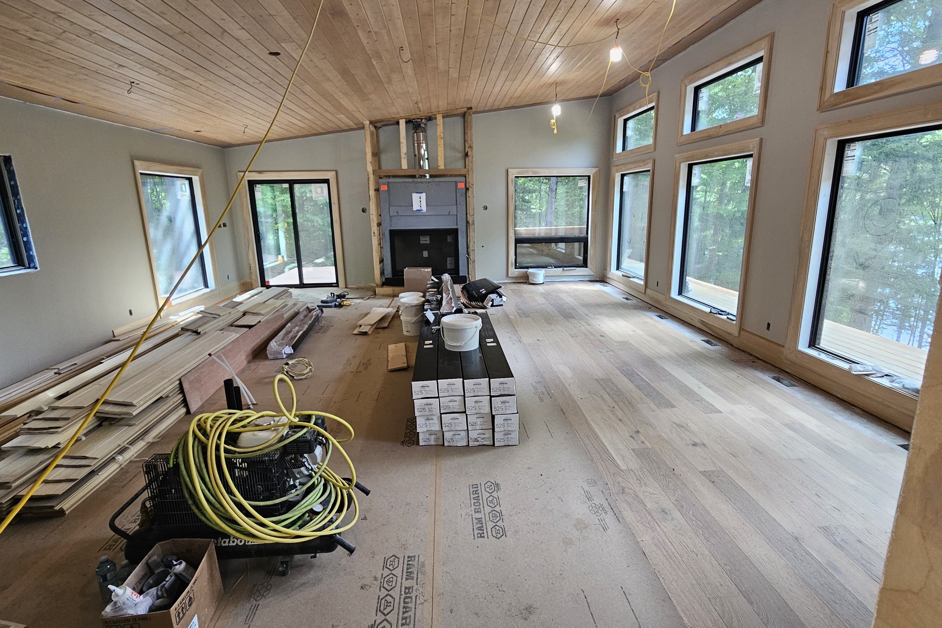 Living Room — Vaulted Wood Ceiling A wide view of a spacious living room under construction, featuring a wooden ceiling, large windows, and new hardwood flooring being installed.