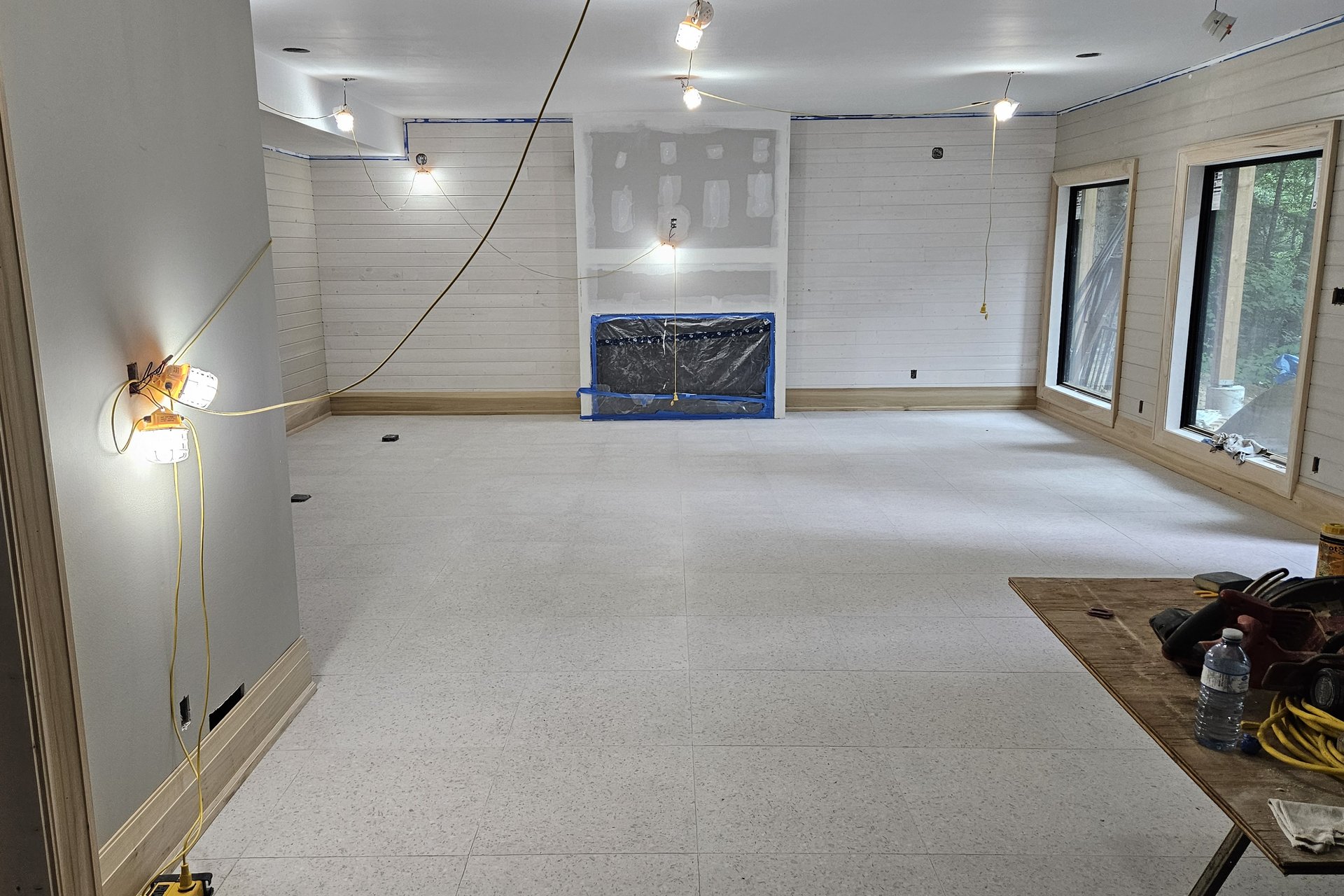 Living Room — Fireplace A wide interior shot of a spacious room under construction, with partially completed walls, a fireplace covered in blue tape, and temporary lighting.
