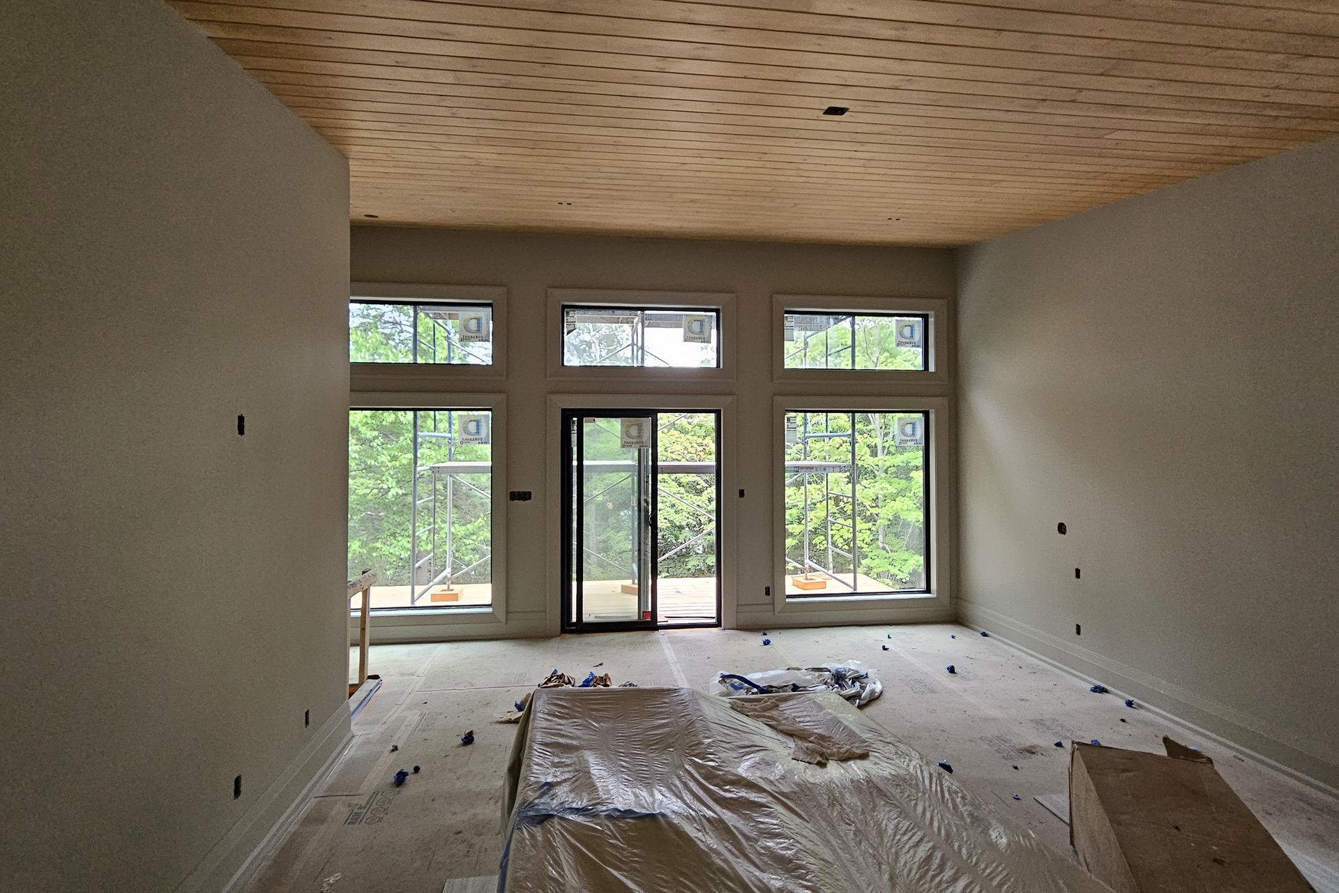 Living Room — Vaulted Wood Ceiling An interior shot of a room with light-colored walls, large windows and a sliding glass door, and a wooden plank ceiling, with construction materials on the floor.