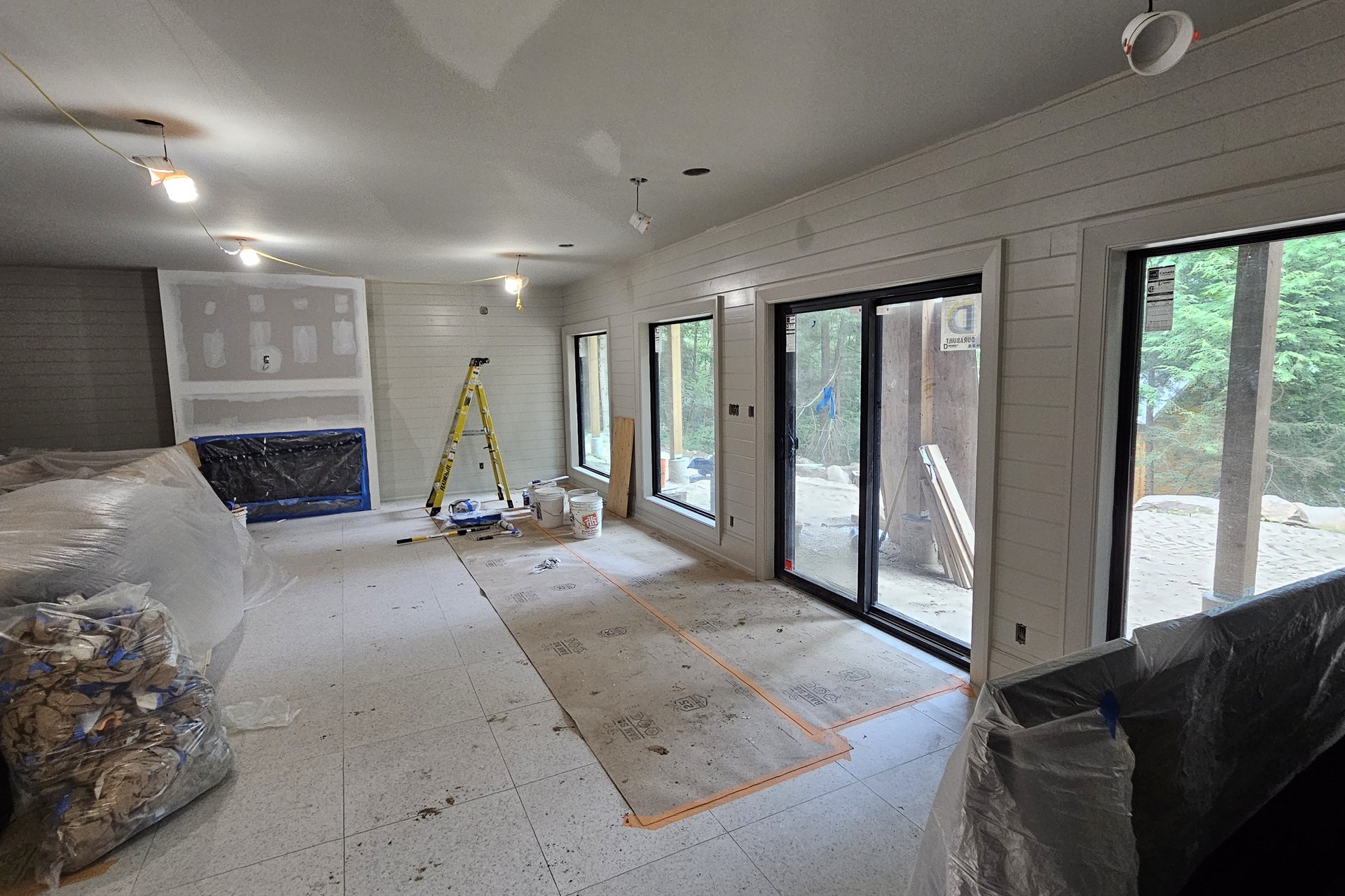 Living Room — Panoramic Windows A spacious living area under construction with shiplap walls, large windows and sliding doors, and protective coverings on the floor and furniture.
