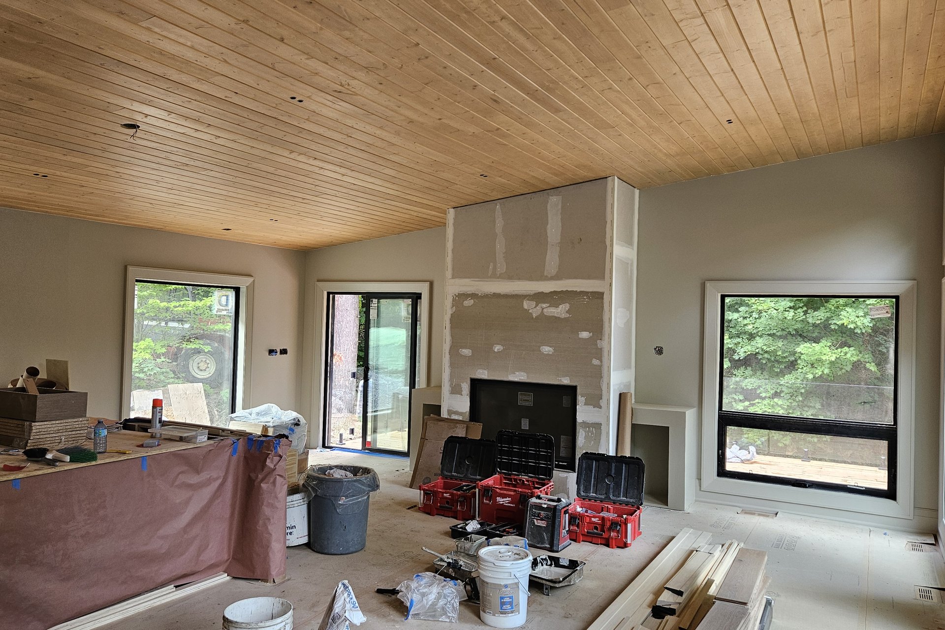 Living Room — Vaulted Wood Ceiling A spacious, unfinished living room features a wooden plank ceiling, light grey walls, a partially covered island, and a fireplace with drywall work, amidst construction tools and materials.