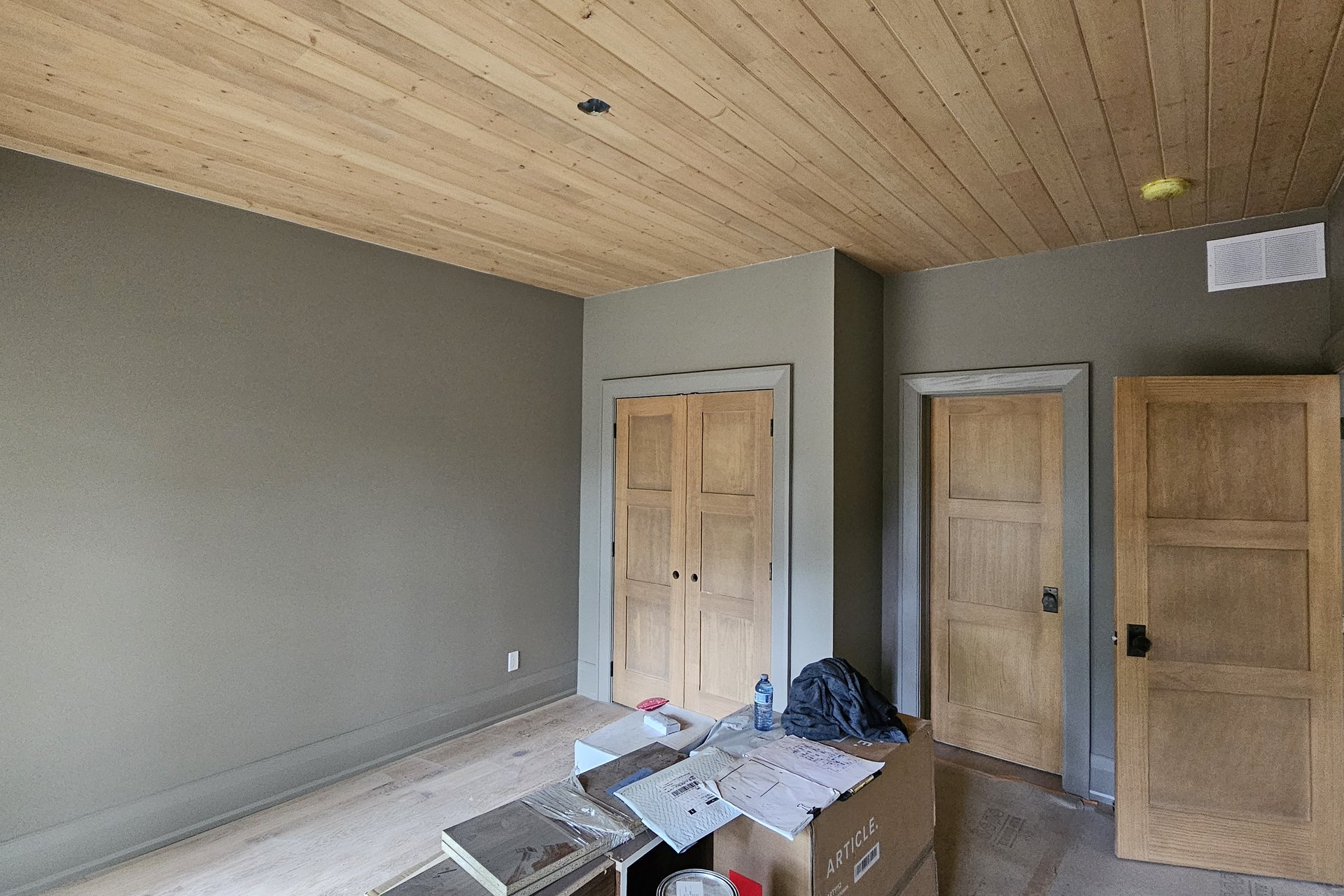 Bedroom — Wood Ceiling A bedroom interior with olive green walls, a light wood plank ceiling, and three wooden doors, with some construction debris on the floor.