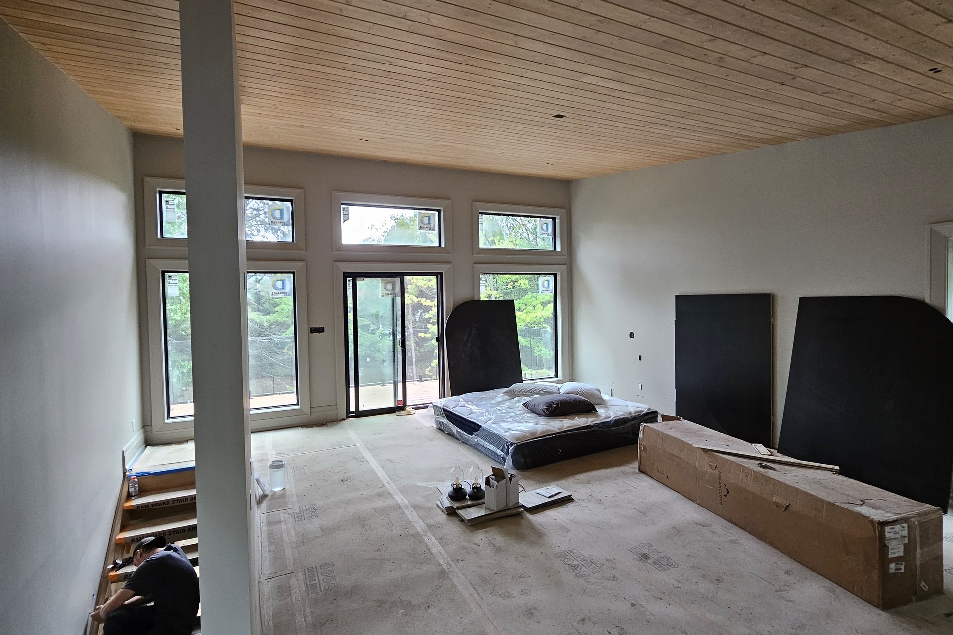 Bedroom — Wood Ceiling A spacious interior room with a finished wooden ceiling, a bed frame, large windows overlooking trees, and construction materials on the floor, seen from an upper landing.