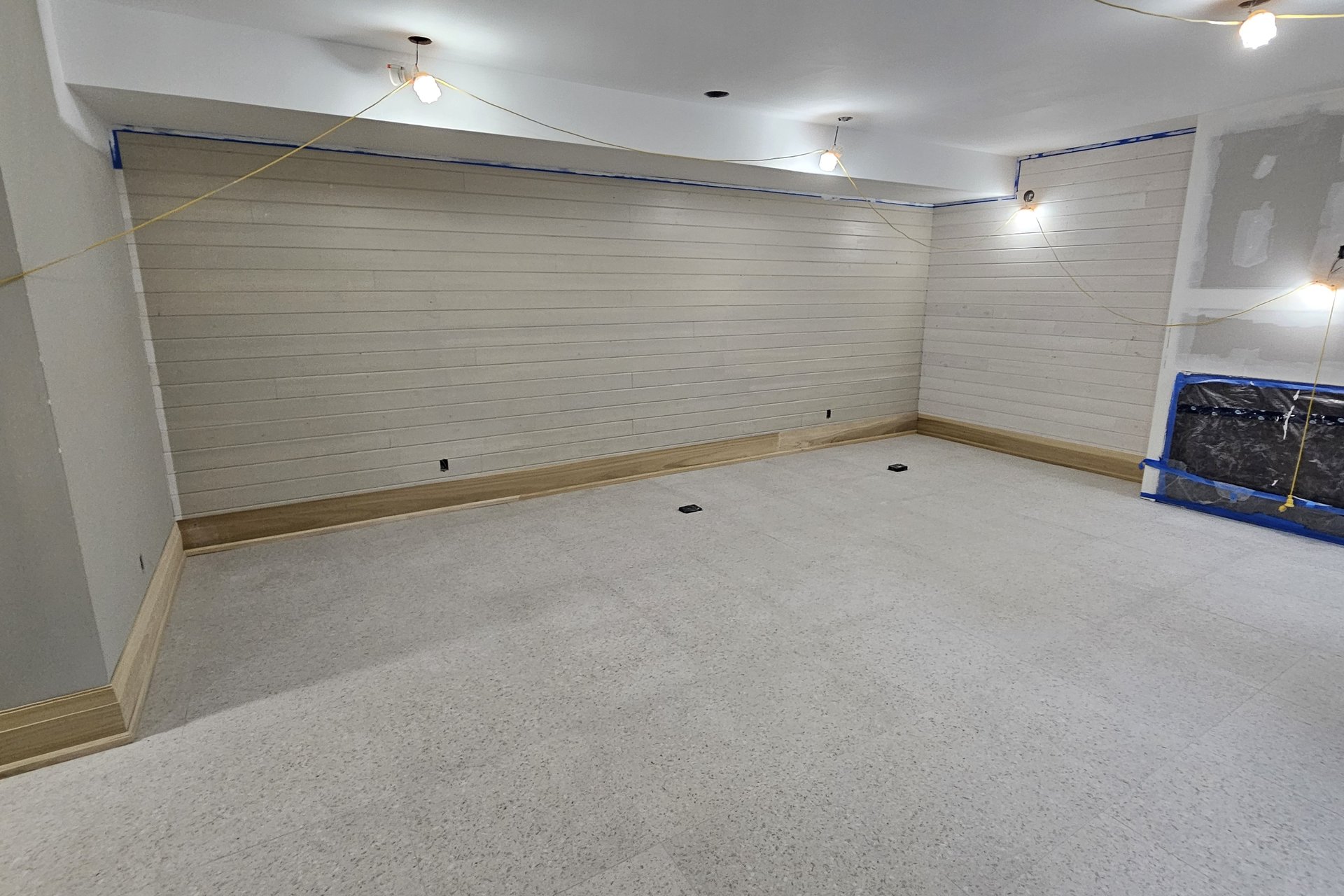 Basement — Finished Space An unfinished basement room features new gray carpet, white painted walls with horizontal paneling, and temporary lighting, with some walls still undergoing drywall finishing.