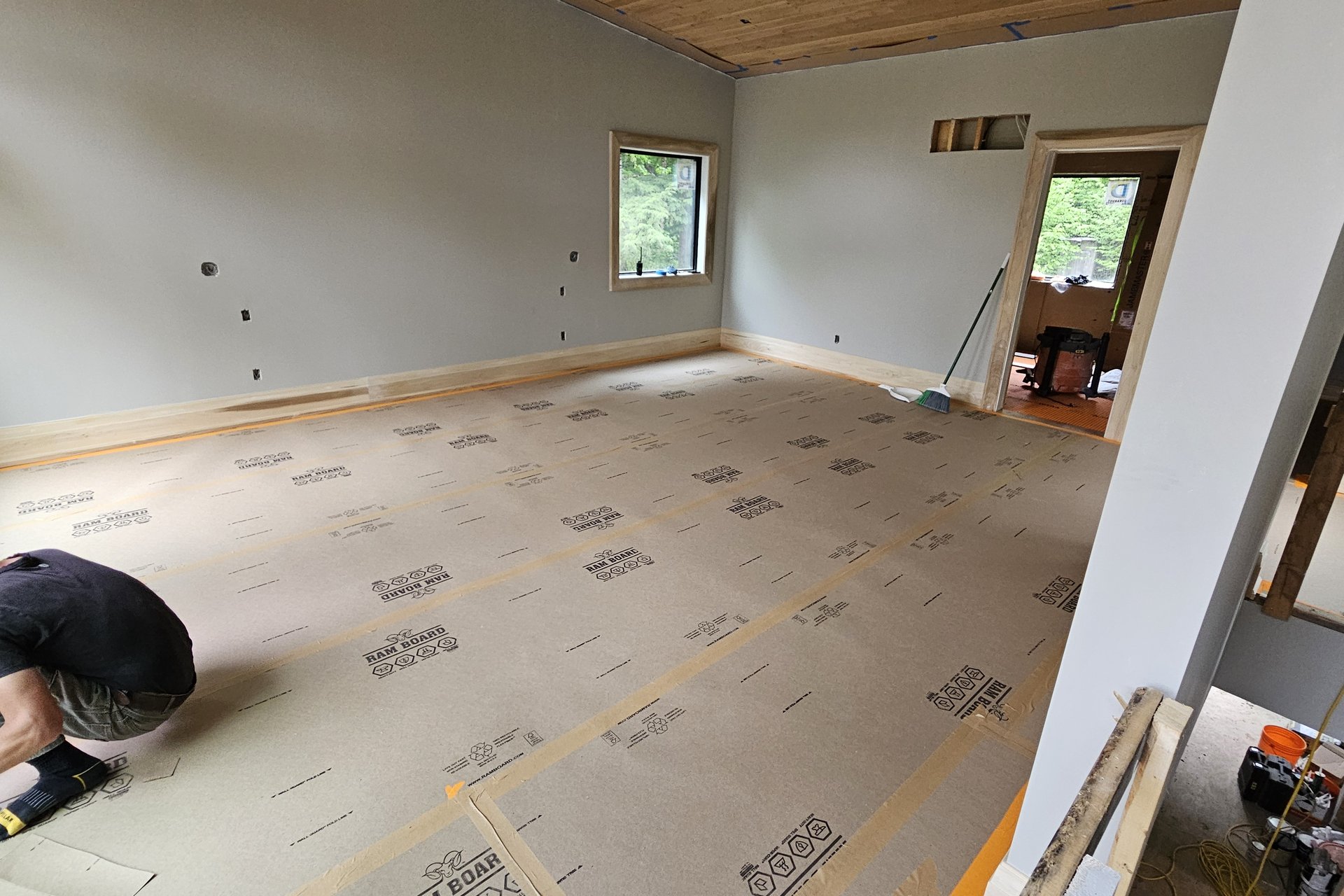 Interior — Subfloor Prep A person is on their knees working on subflooring with insulation laid out in a large, unfinished room with baseboards installed and windows showing trees outside.