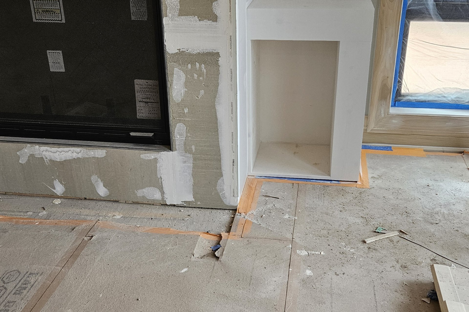 Interior — Finishing The interior of a room undergoing renovation shows a fireplace area with unfinished drywall, a small built-in cubby, and a window covered for protection.