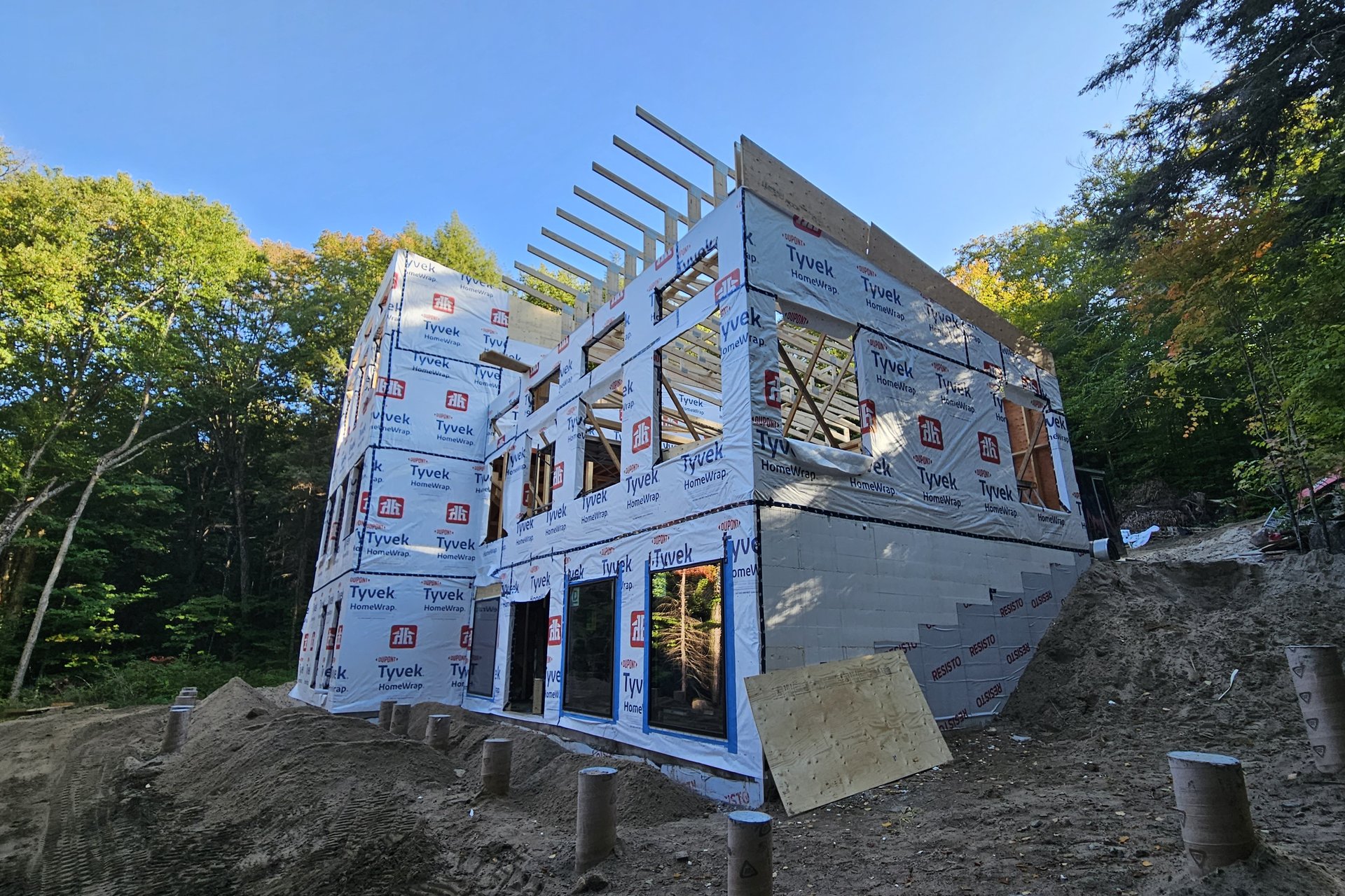 Construction — Roof Framing A large house frame wrapped in Tyvek, with windows installed and exposed rafters on the roof, surrounded by trees.
