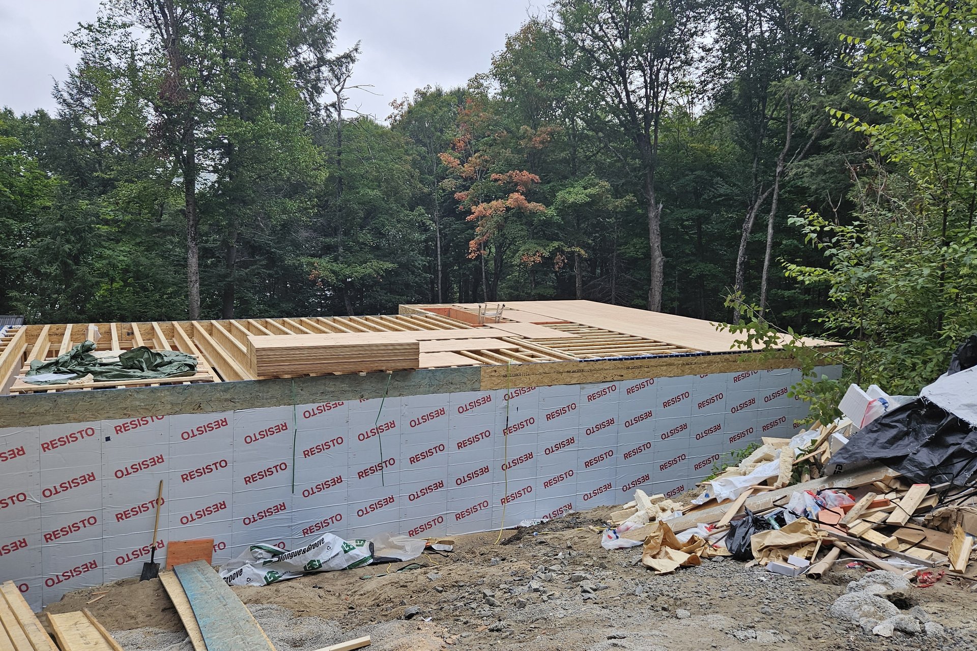 Construction — Framing Stage An exterior view of a building under construction shows the floor framing and insulation on the foundation walls, with construction debris piled nearby and trees in the background.
