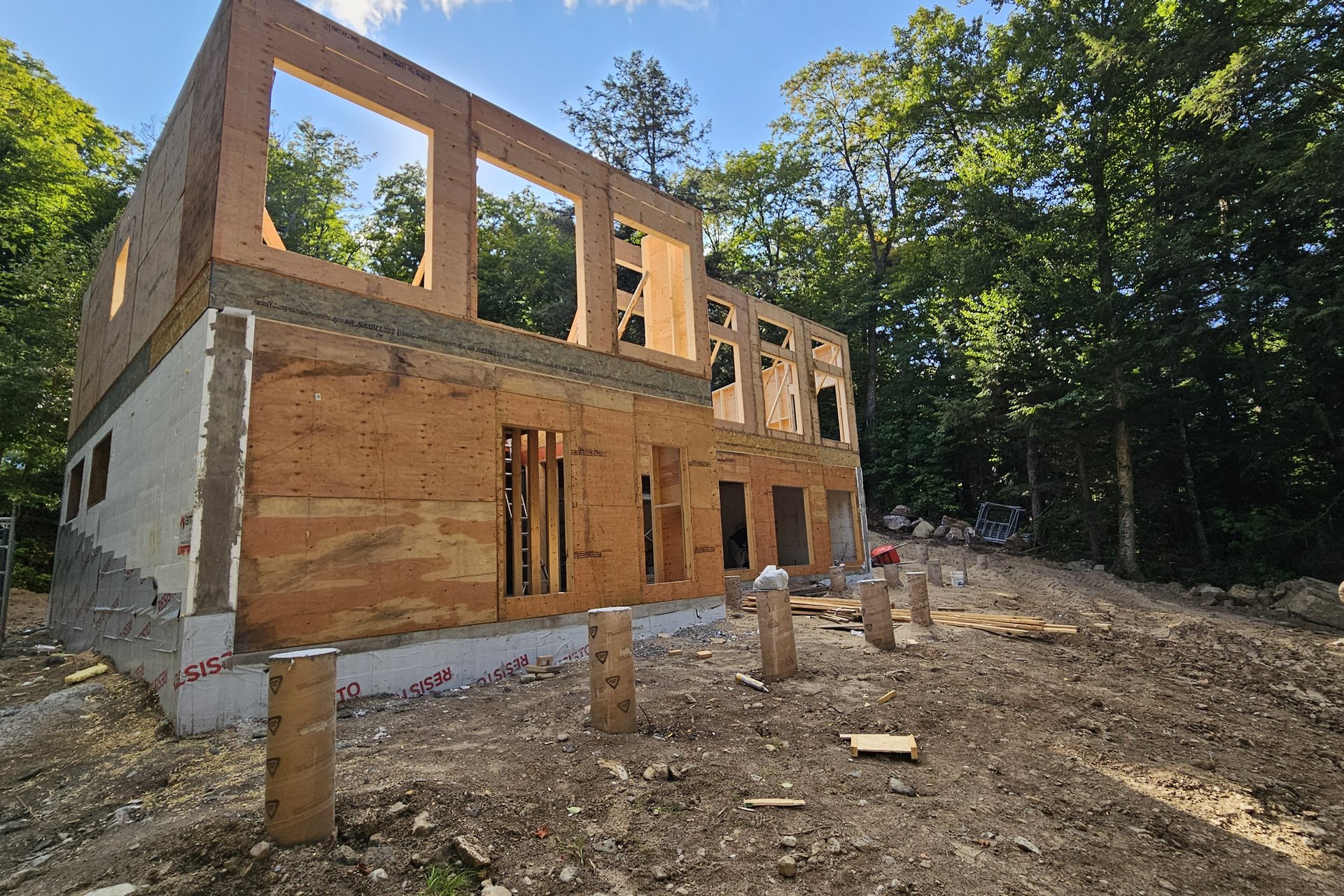 Construction — Framing Stage A house under construction shows plywood sheathing partially covering the framed structure on a sunny day, with a dirt foreground and surrounding trees.
