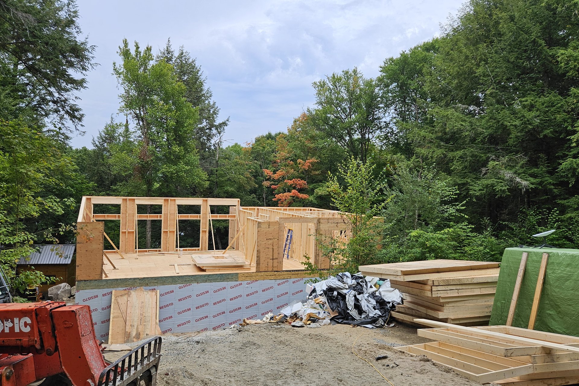 Construction — Framing Stage An exterior view of a house under construction, demonstrating the framing stage with timber walls and some windows installed, surrounded by a wooded area.