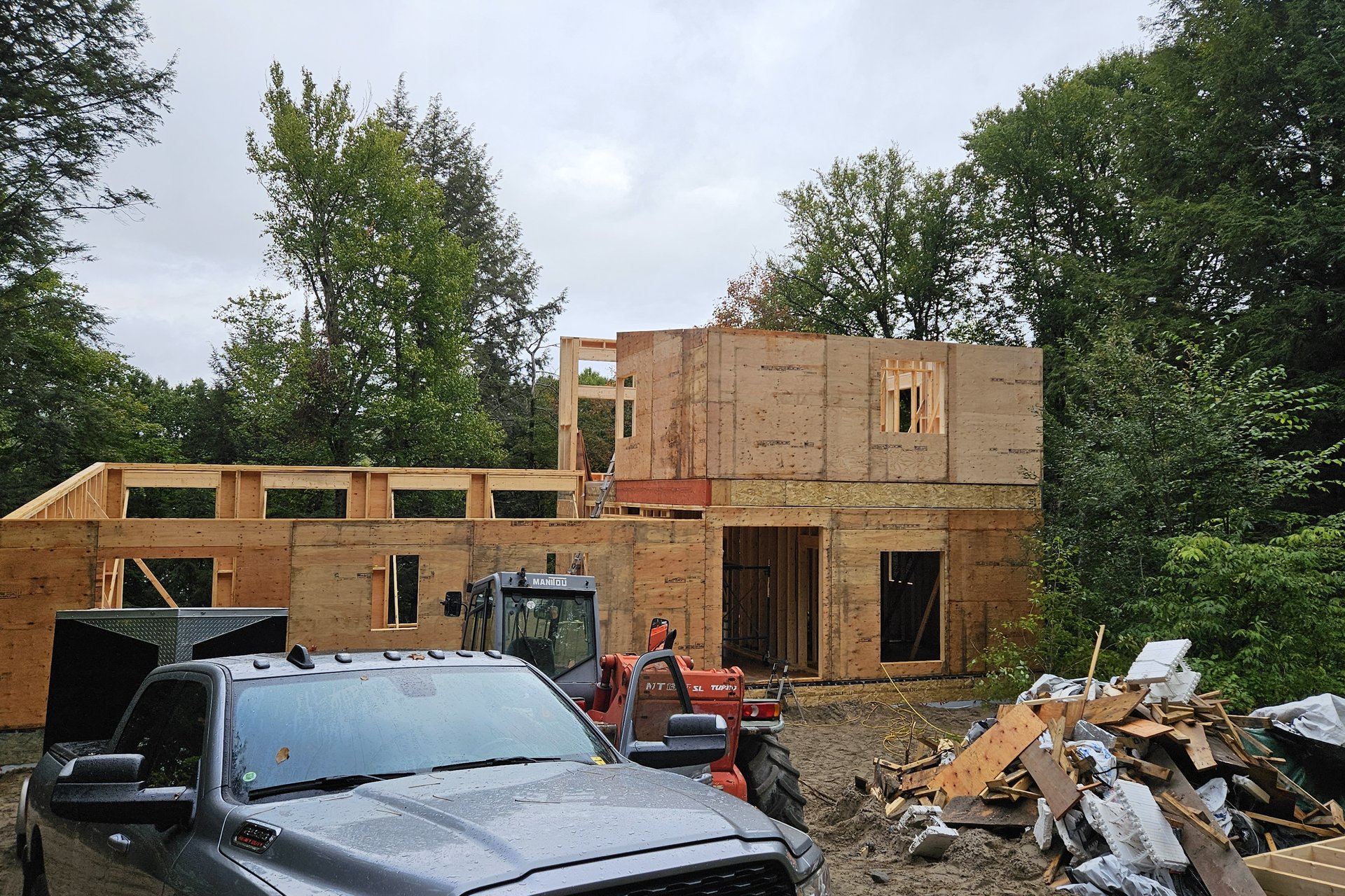 Construction — Framing Stage A partially framed house with plywood sheathing is visible behind a pickup truck and a pile of construction debris, surrounded by trees under an overcast sky.