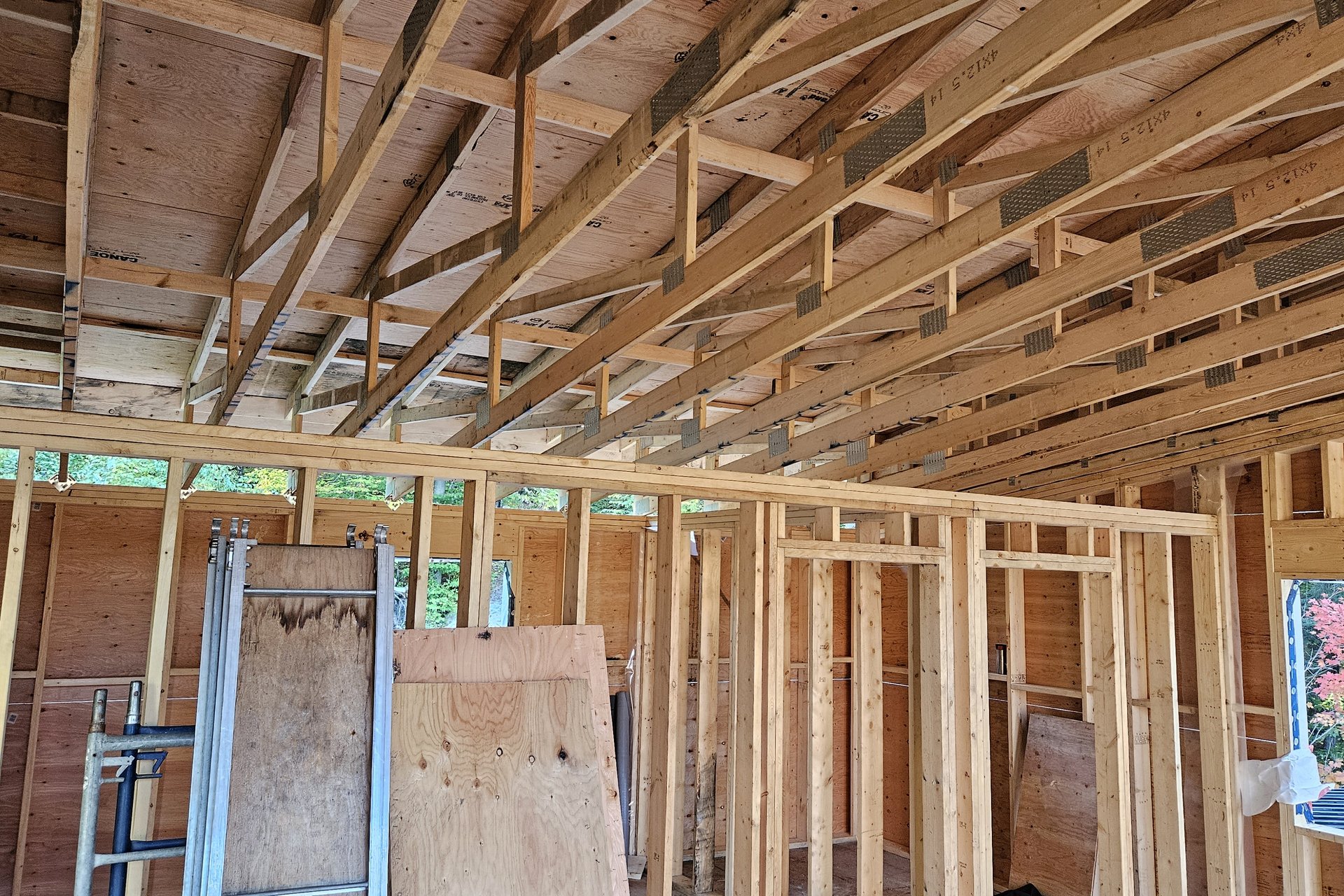 Construction — Interior Framing An interior view of a house frame under construction, showing wooden wall studs, ceiling joists, and a partial view of a window overlooking colorful autumn trees.