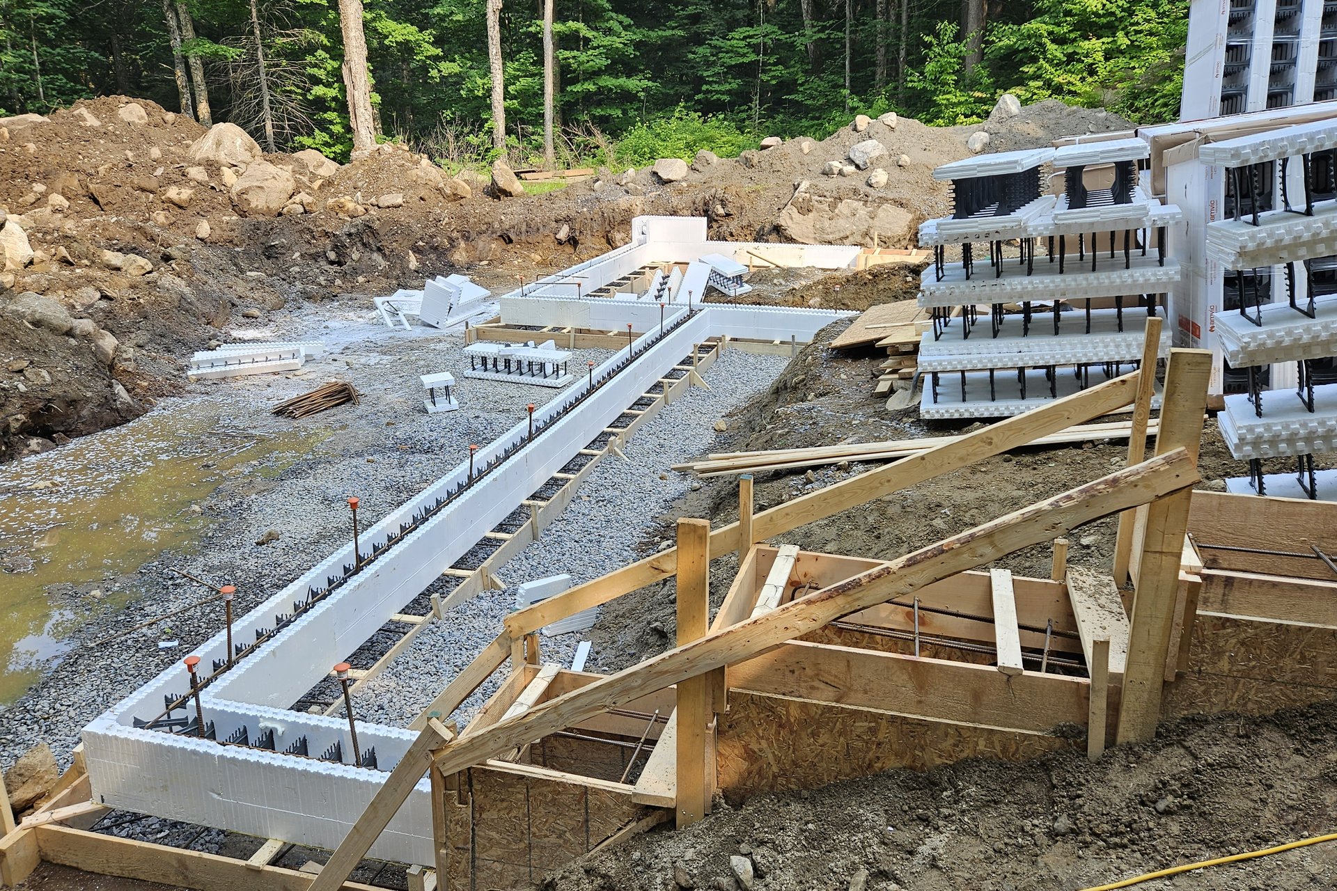 Foundation — Formwork The foundation forms for a new building are visible on a cleared lot, with various building materials stacked nearby and wooded areas in the background.