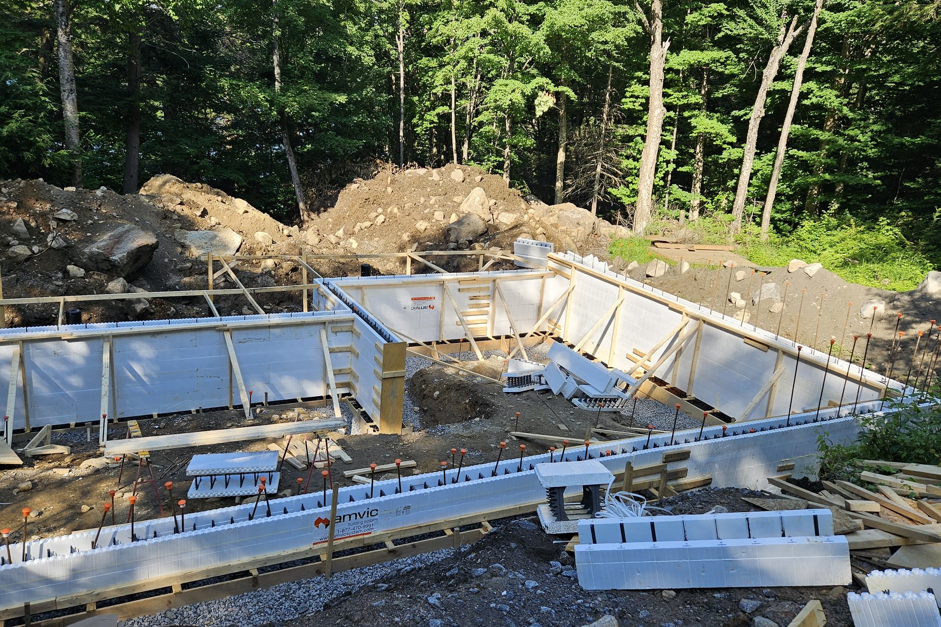 Foundation — Aerial View An aerial view of the foundation construction site, showing several sections of the concrete foundation walls and surrounding earthwork with trees in the background.