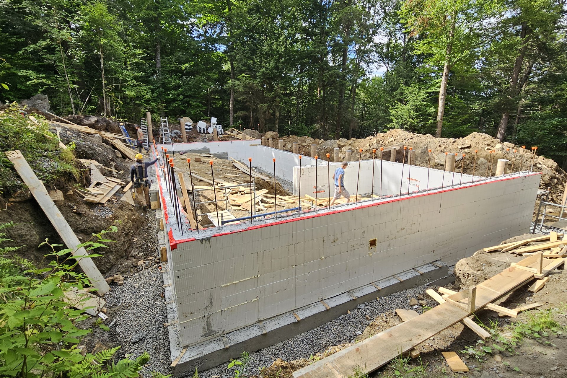 Foundation — Aerial View An aerial-perspective image captured from atop a rocky outcrop showing the foundation of a house under construction with worker scaffolding and lumber piles.