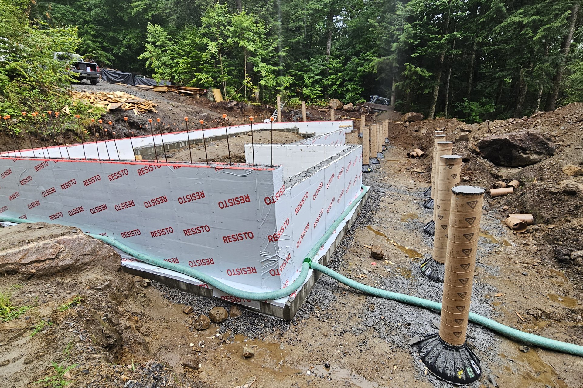 Foundation — Formwork The foundation walls of a house are visible, covered with insulating material, with green pipes running around the base and cylindrical pier forms standing nearby.