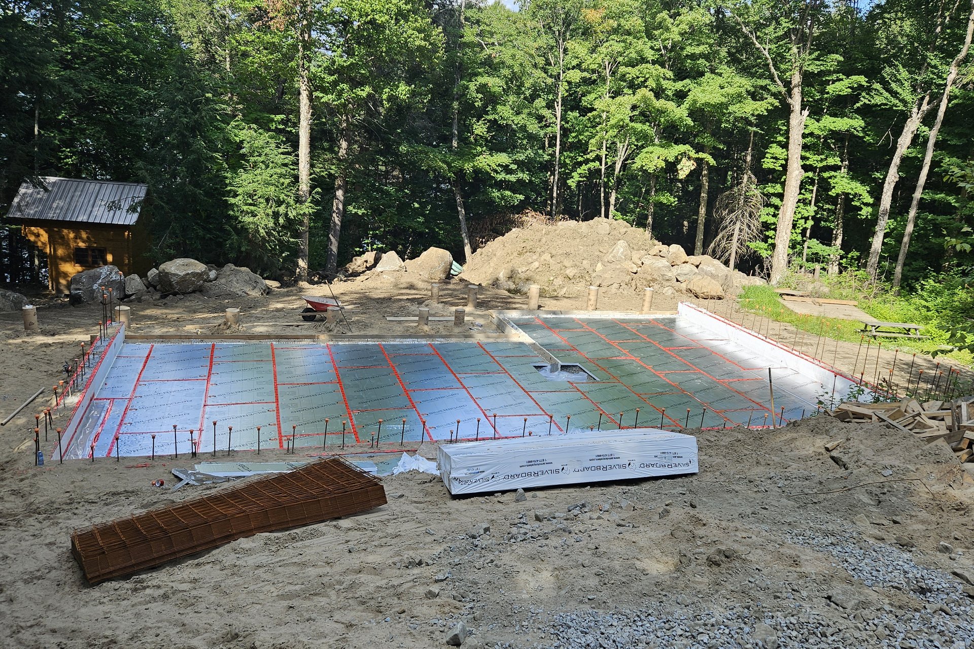 Foundation — Aerial View An aerial view of a construction site showing a foundation with radiant heating pipes laid out on insulation boards, surrounded by excavated dirt and trees.