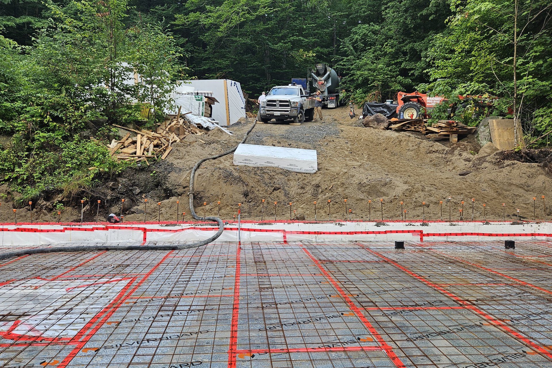 Foundation Work The foundation area is being prepared for concrete with red radiant heating tubes laid out on a grid of rebar, and a concrete truck visible in the background.
