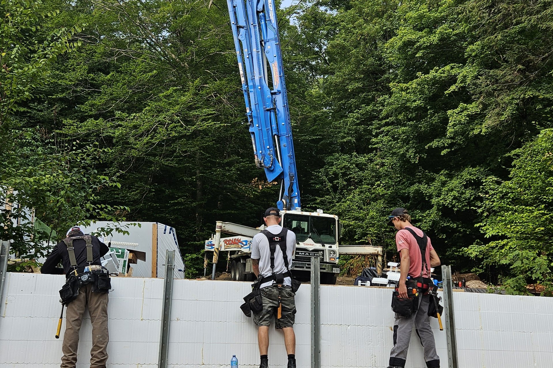 Foundation — Concrete Pour Three construction workers stand on scaffolding, working on a foundation wall made of white insulating concrete forms, with a concrete pump truck in the background.