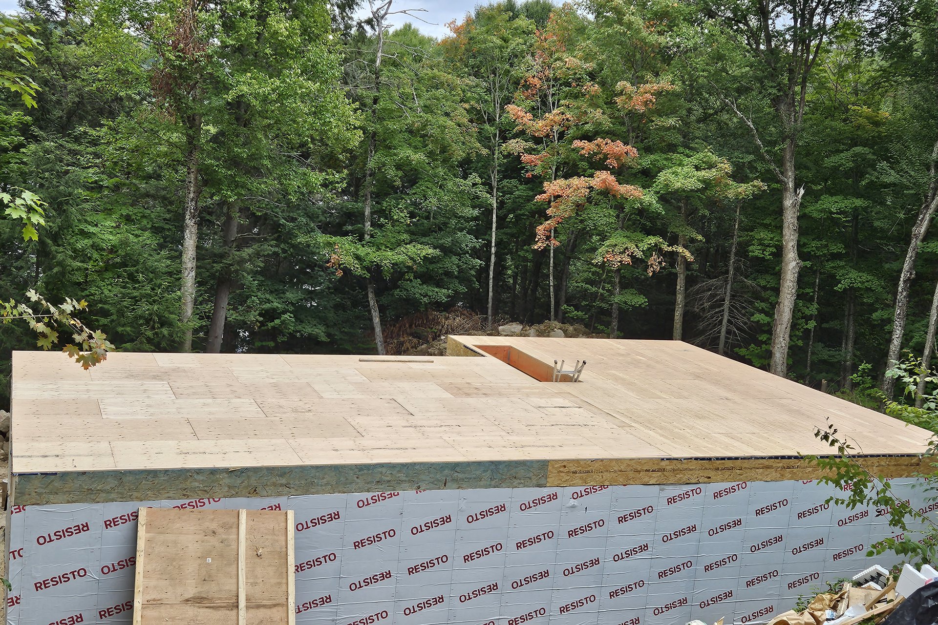 Roofing An aerial view of a new building's roof under construction, featuring wooden sheathing and green weather-resistant barrier on the walls, surrounded by lush trees.