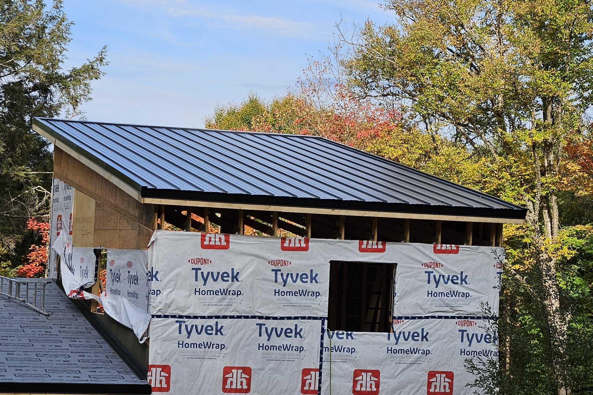 Roofing — Metal Roof Installation An exterior view of a house addition under construction, showing a newly installed metal roof, framed walls wrapped in Tyvek, and surrounding autumn trees.