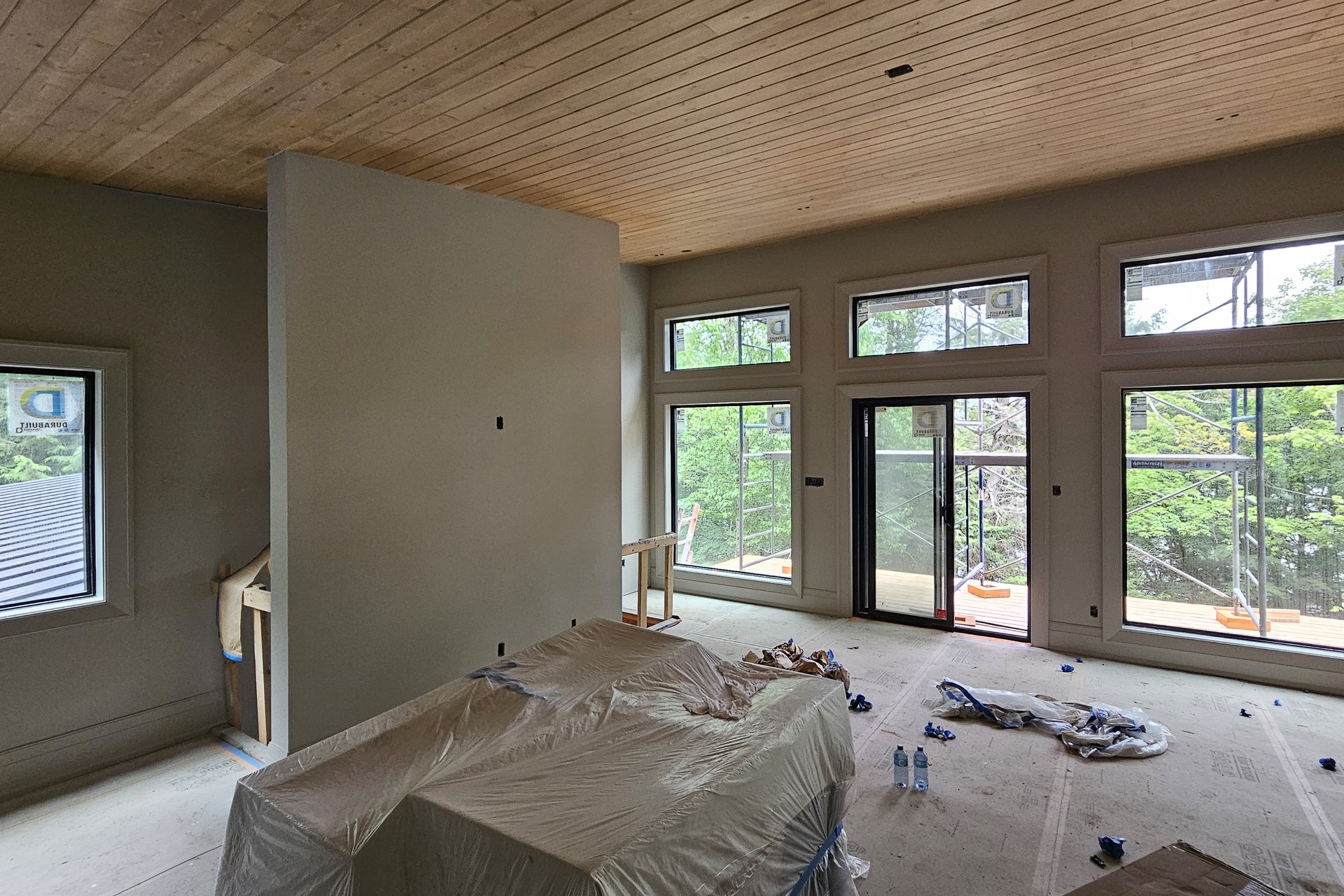 Construction An interior room under construction featuring a wood panel ceiling, light green painted walls contrasting with white unfinished walls, and large windows looking out onto trees.