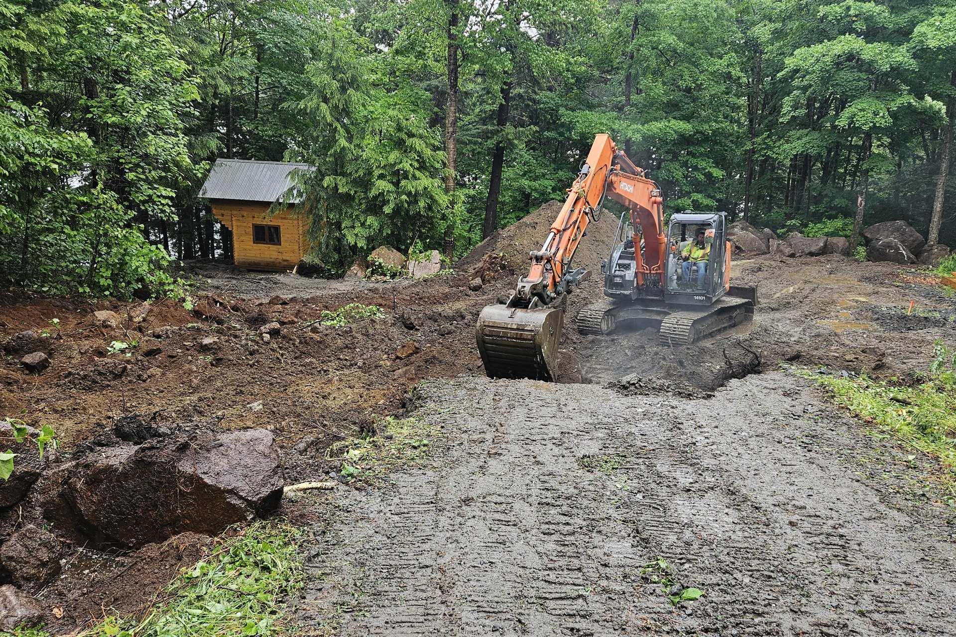 Woodland Landscape An excavator is digging and moving dirt on a muddy construction site surrounded by trees, with a small wooden structure in the background.