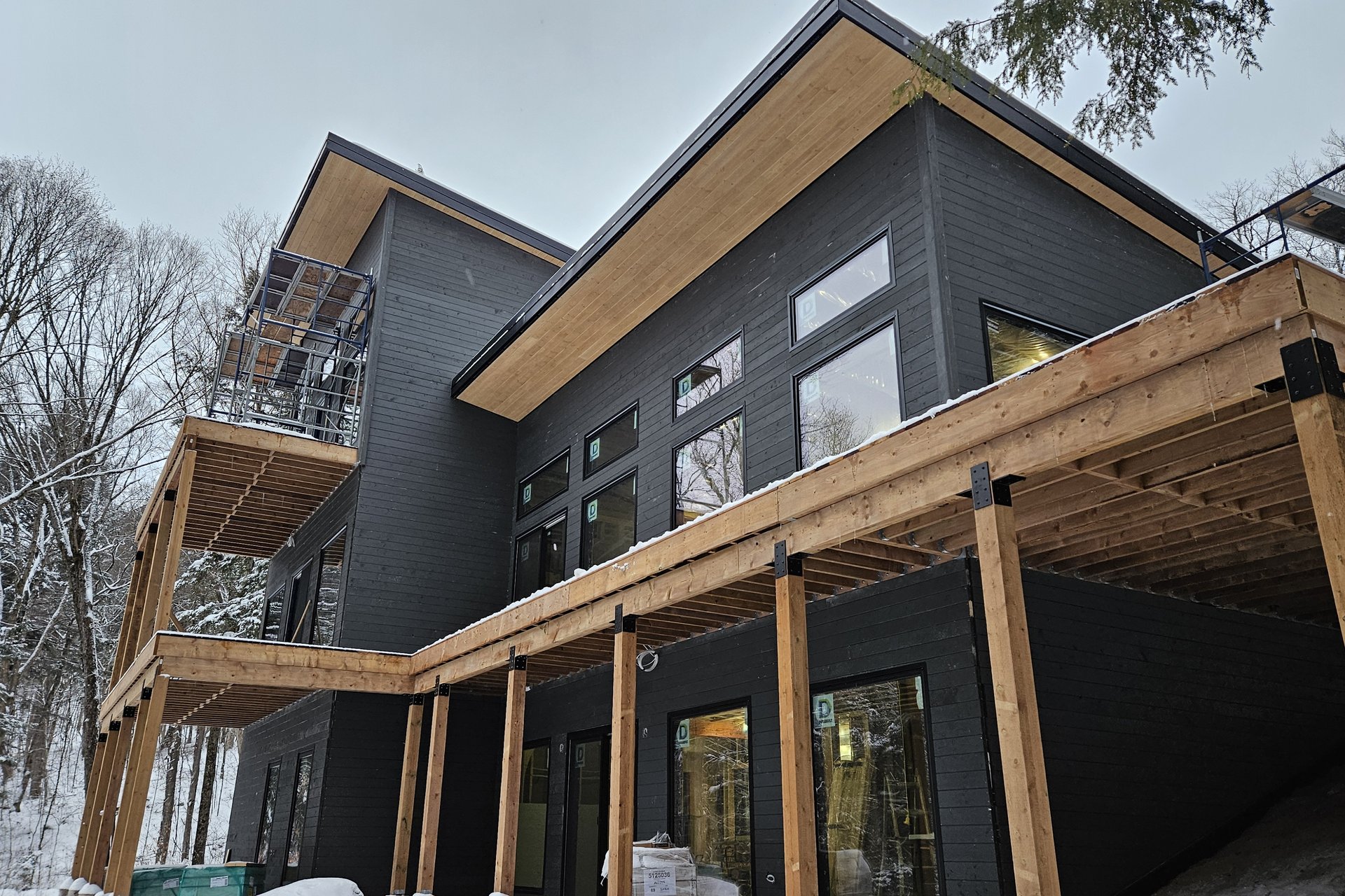 Exterior — Siding Installation An exterior view of a modern house under construction with dark siding, large windows, and wooden eaves, surrounded by a snowy landscape.