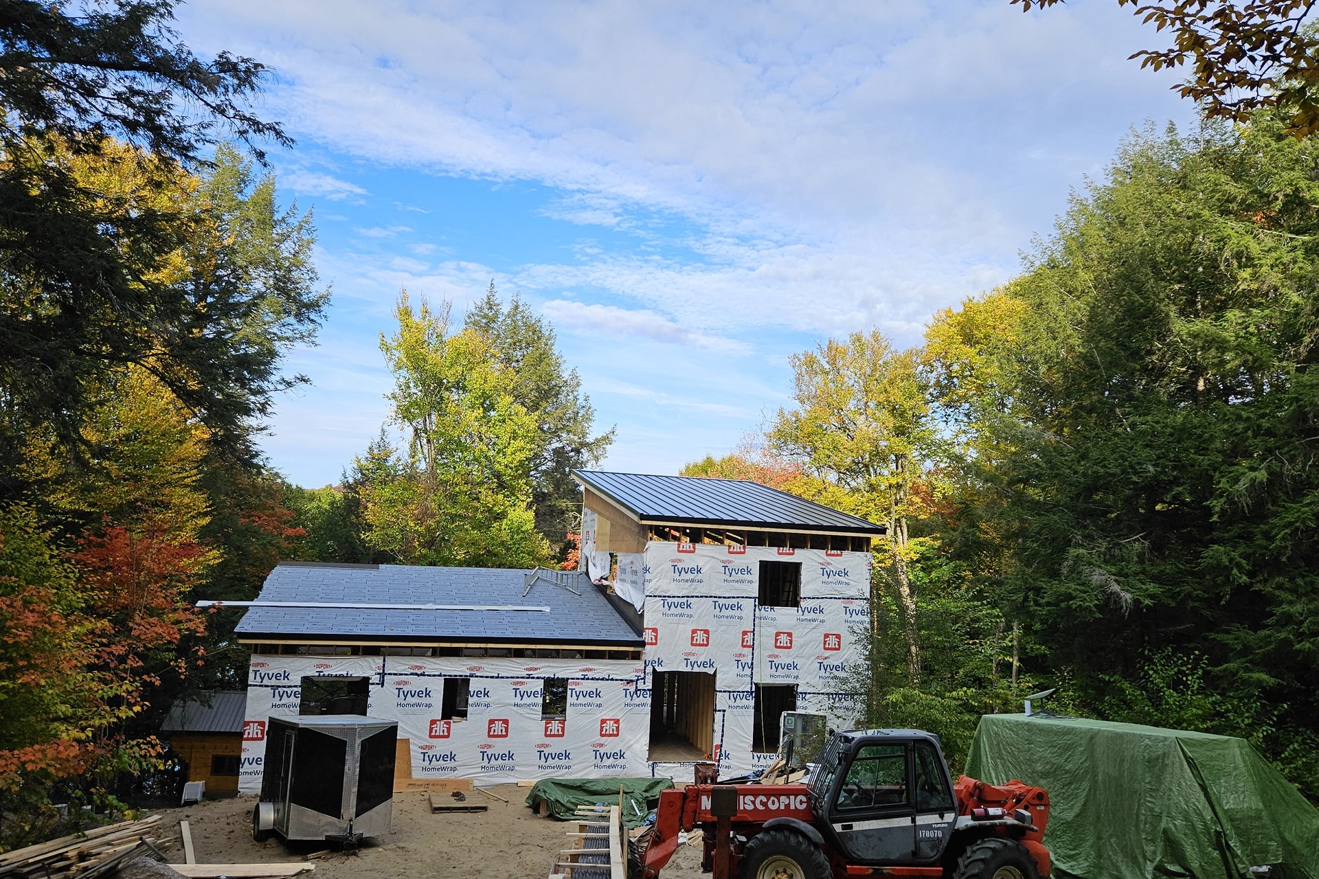 Exterior — Roof Progress An exterior view of a house under construction with a dark metal roof, wrapped in white sheathing, surrounded by lush green and autumnal trees under a partly cloudy sky.