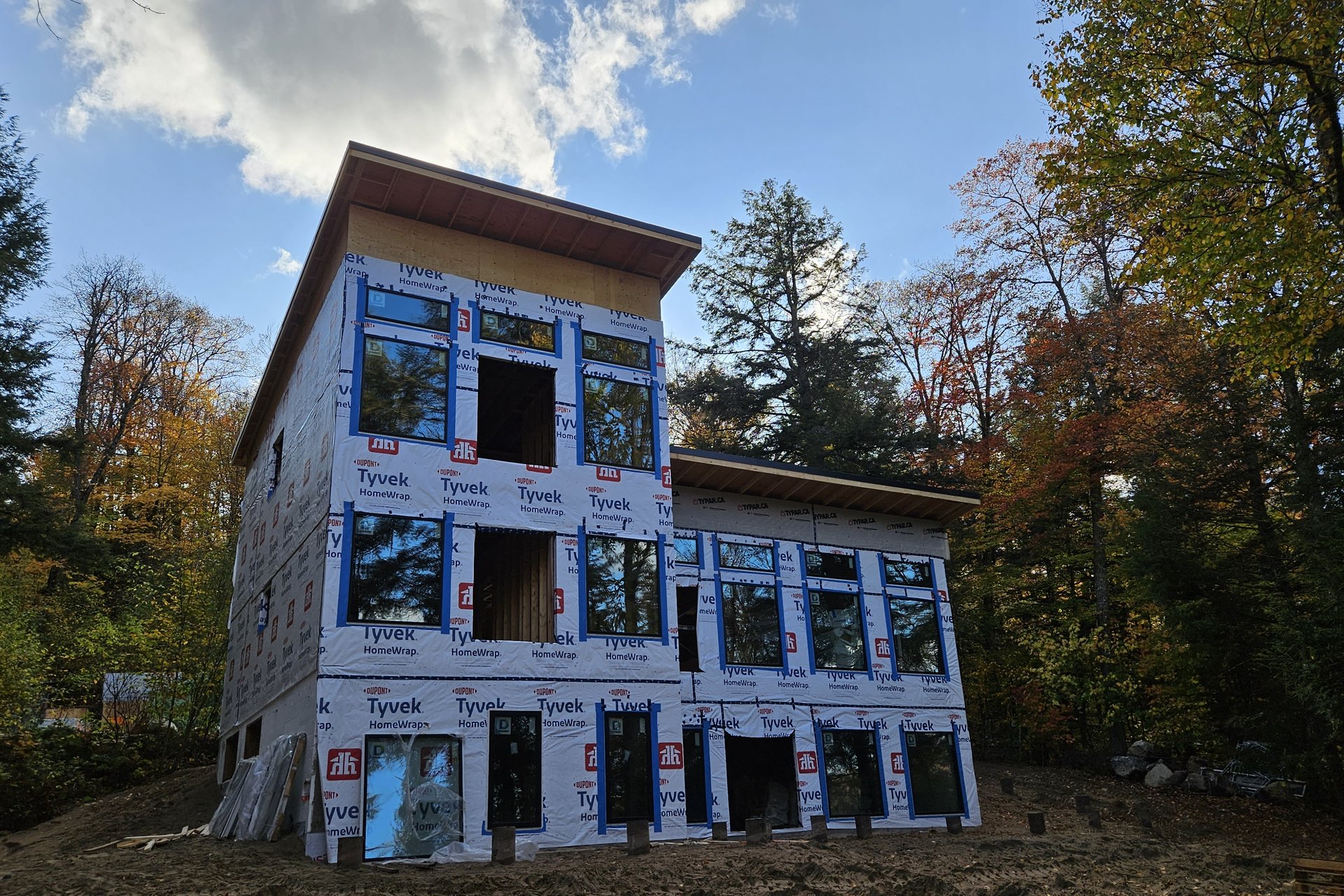 Exterior — Window Installation A modern, multi-story house clad in Tyvek wrap and featuring numerous large windows, set against a backdrop of trees with some autumn foliage under a blue sky.