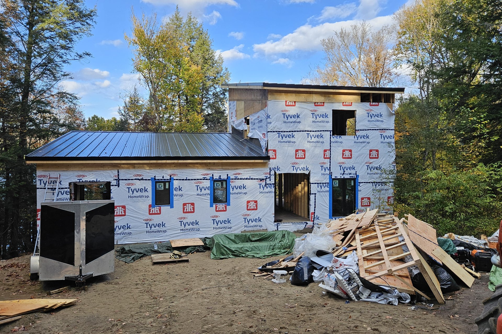 Exterior — Roof Progress An exterior view of a house under construction, with Tyvek wrap on the walls, a finished black metal roof on one section, and construction debris on the ground.