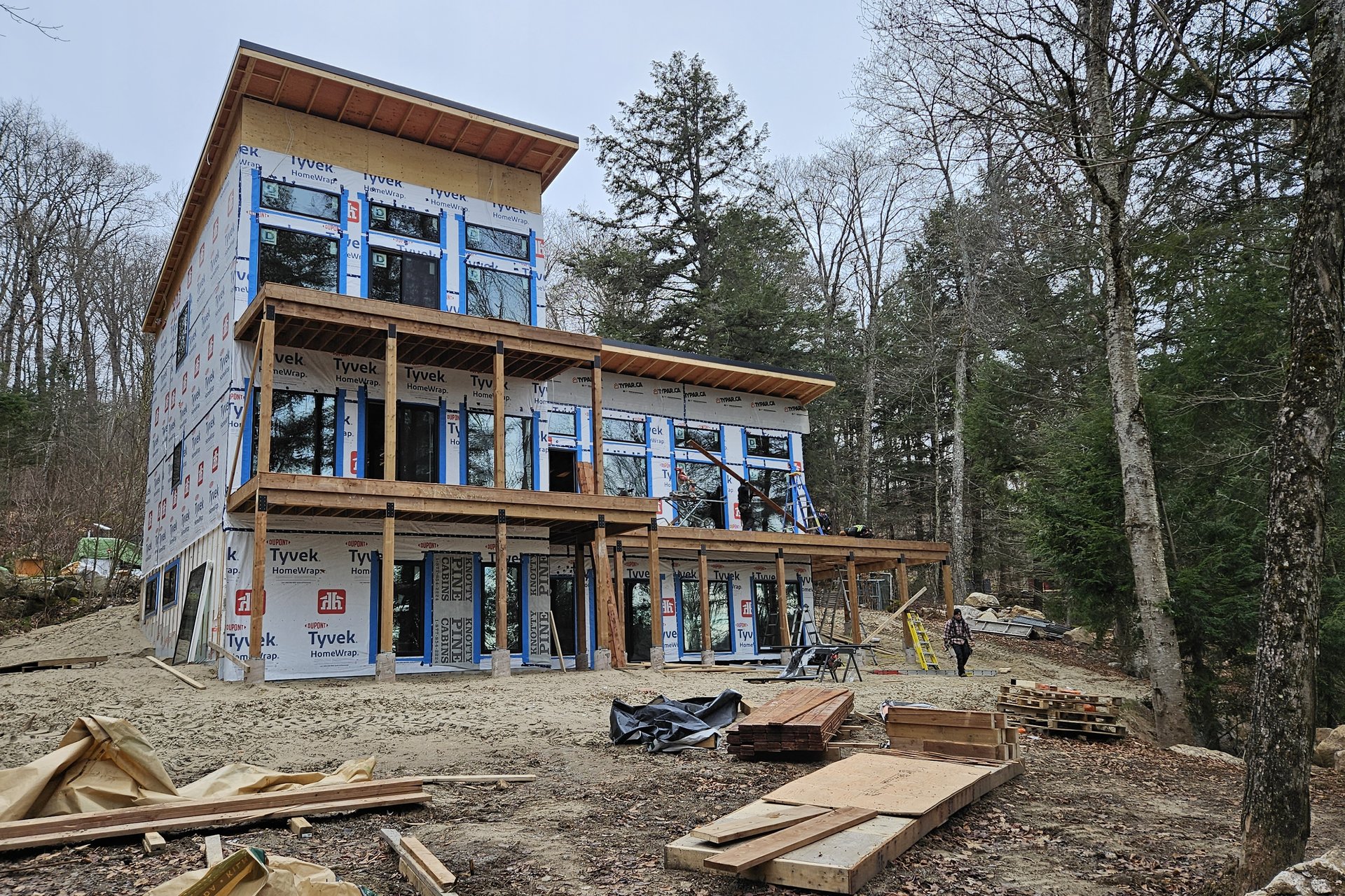 Exterior — Window Installation A two-story house under construction features large windows, exposed wooden framing for balconies, and a dirt ground surrounding it with trees in the background.