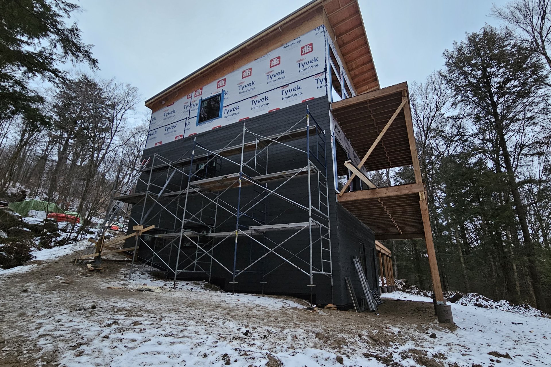Exterior — Deck Construction A multistory house under construction is covered in black house wrap, featuring scaffolding on one side and a wooden deck frame on the other, set against a snowy, wooded background.