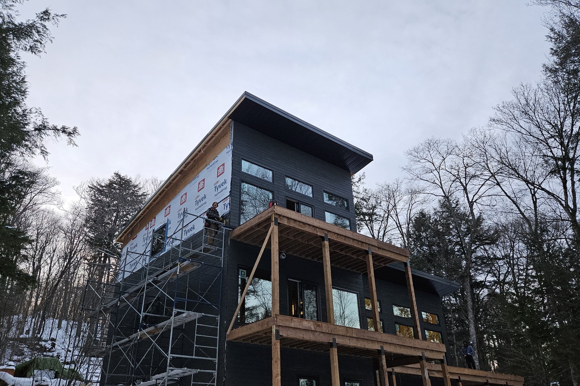 Exterior — Siding Installation A modern multi-story house under construction with black siding, wrapped sections, scaffolding, and wooden balconies, surrounded by trees and a snowy ground.