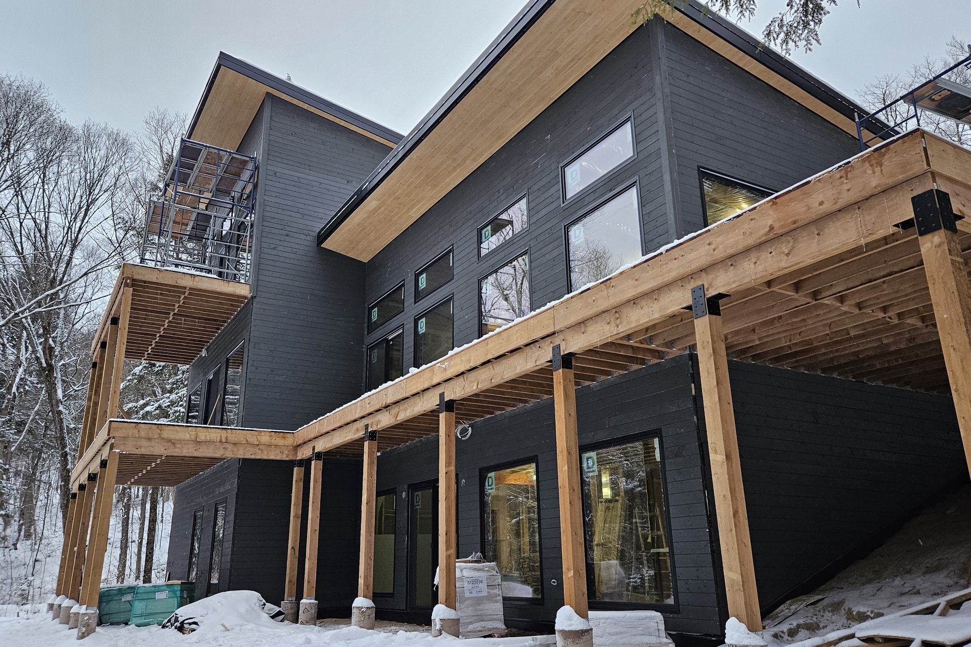 Exterior — Siding Installation A modern house with dark siding, large windows, and extensive wooden deck structures is shown amidst a snowy, wooded landscape, still under construction.
