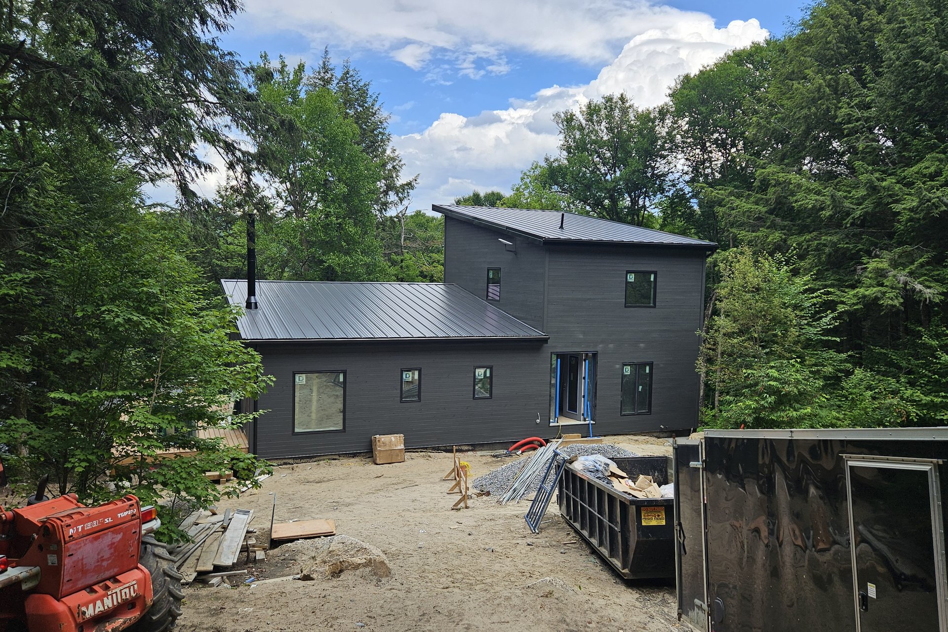 Exterior — Roof Progress A modern black house with a dark metal roof is under construction, surrounded by trees, with a construction dumpster and equipment visible in the foreground.