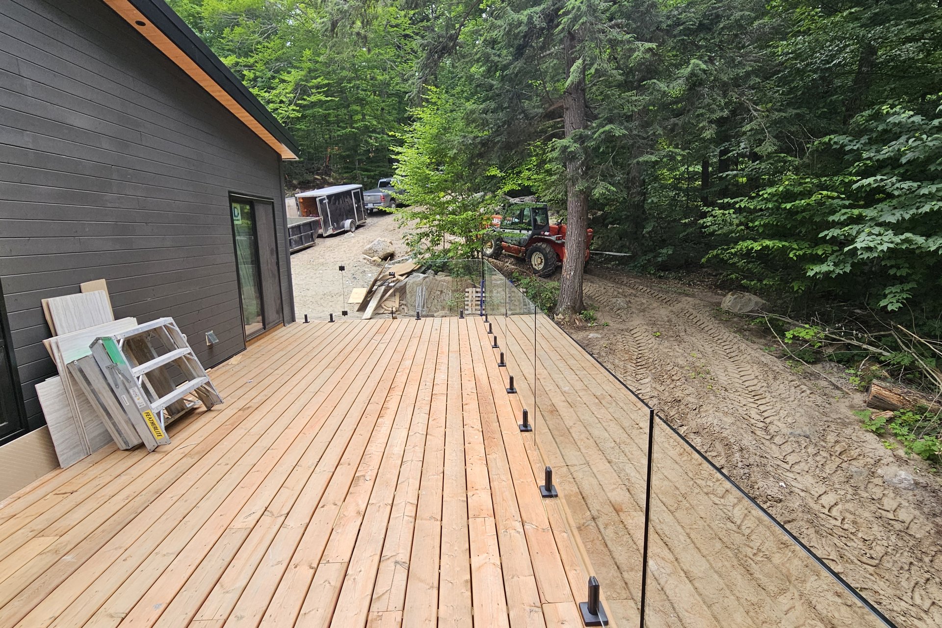 Exterior — Deck Construction An outdoor wooden deck with glass railings is shown next to a modern black house, with construction materials and a tractor visible in the background, suggesting ongoing work.